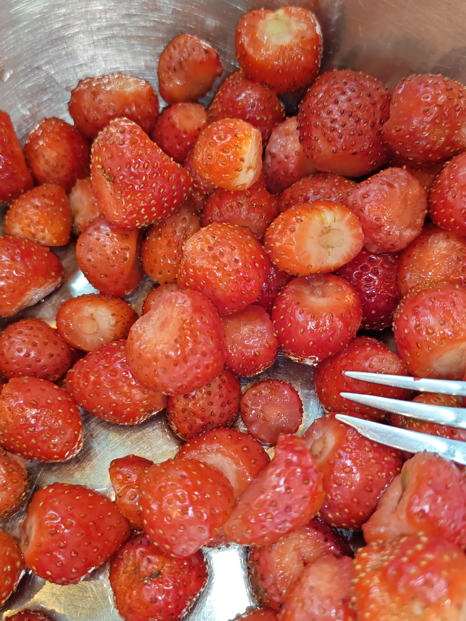 A close-up, overhead shot shows a pile of halved strawberries in a metallic bowl. The strawberries are various shades of red, with small yellow seeds visible on their surfaces. Some strawberries appear to be whole, while others are cut in half, revealing their white interiors. The bowl's metallic surface is visible around the edges of the fruit. In the bottom right corner of the image, a portion of two silver forks is visible, with their tines resting on top of some of the strawberries. The scene is lit from above, casting soft shadows and highlighting the glistening texture of the fruit.