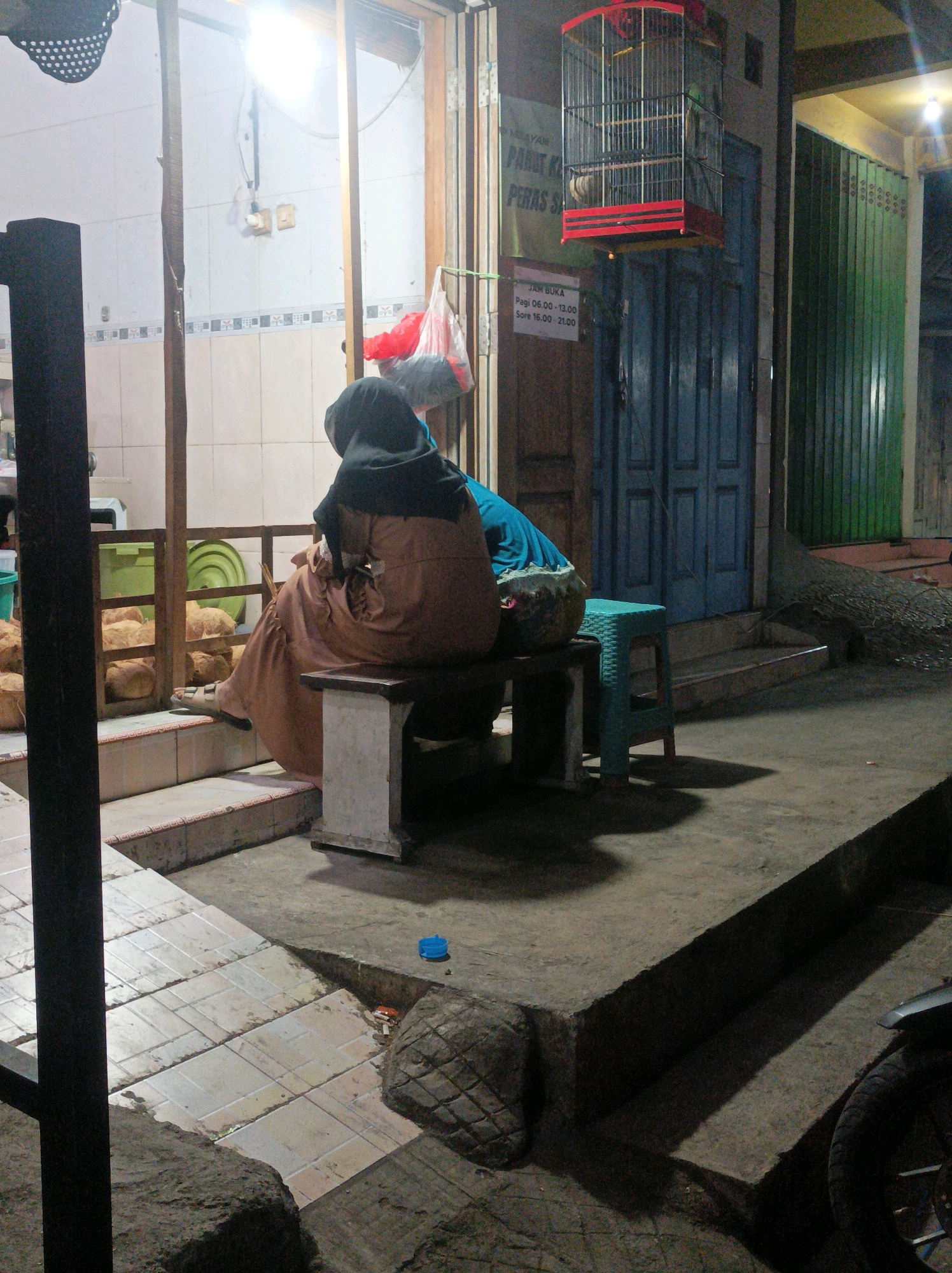In the city of Sumberagung, Indonesia, at dusk, two people are sitting on a bench outside a shop. The person on the left, wearing a black hijab and a brown robe, is facing away from the camera. Their legs are crossed, and one foot, wearing a sandal, rests on the tiled step. The person on the right is mostly obscured, but a portion of their head and shoulders, draped in a blue cloth, is visible. They appear to be leaning forward.
Behind them, the shop's interior is lit by overhead lights, revealing tiled walls and a counter. A birdcage hangs from the ceiling. In front of the shop, stacked on shelves, are several round, light-brown objects, possibly loaves of bread or coconuts. A teal plastic stool stands beside the bench.
The ground in front of the shop is paved with square tiles, and a concrete step leads up to the shop entrance. A small blue plastic cup is on the ground near the step. To the right of the frame, a portion of a motorcycle is visible, including its wheel and part of the frame. The overall scene is dimly lit, suggesting either early morning or late evening.
