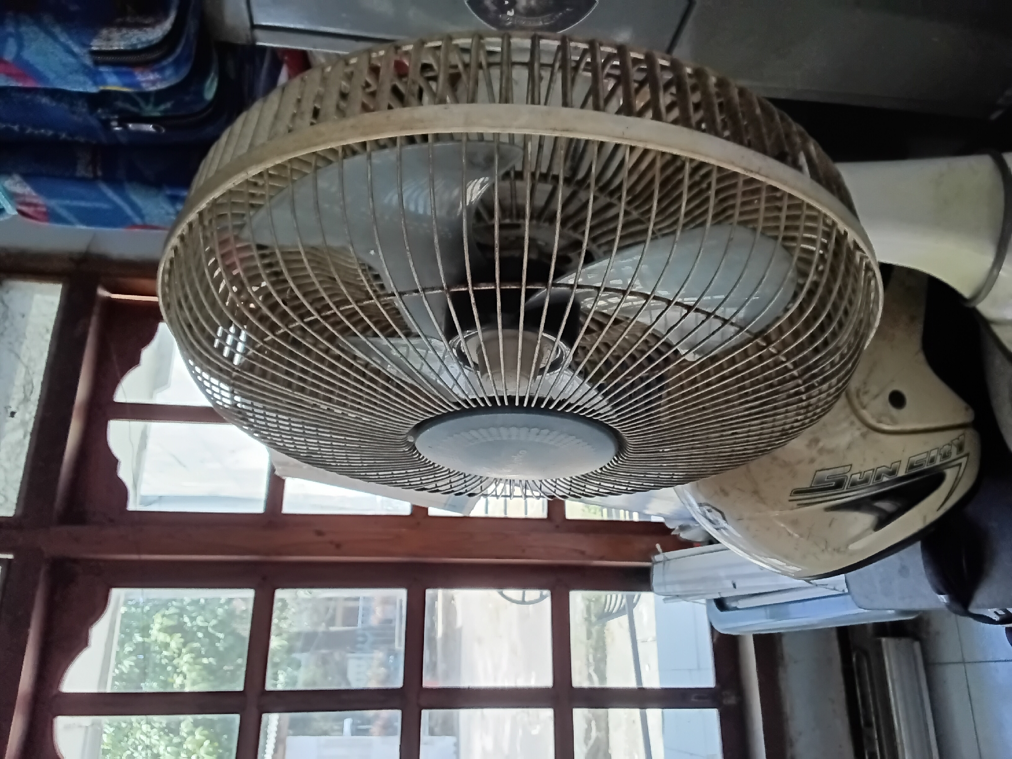 A white and beige electric fan is positioned on top of a white and black helmet. The fan is made of a beige metal grill with a white center piece. The helmet is white with black and silver markings. In the background, a wooden framed window with multiple panes shows greenery and buildings outside. To the right of the fan, a portion of a blue and red patterned bag is visible. The scene appears to be indoors.