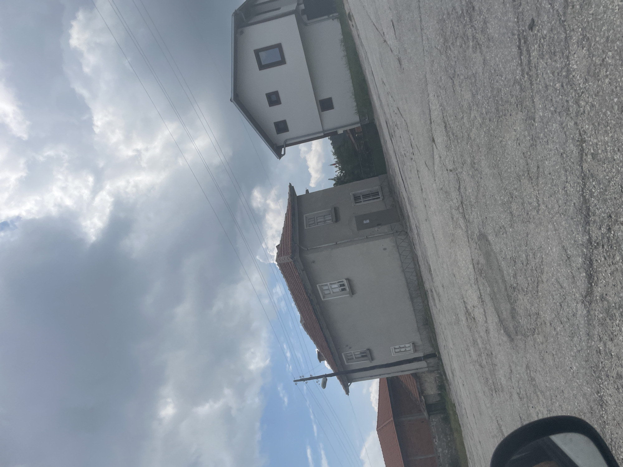 The image depicts a view of a street in the city of Dobrostan, Bulgaria, on a cloudy day. In the foreground, the road surface is cracked asphalt. To the left, a portion of a car's side mirror is visible. In the middle ground, two houses are present. The house on the left is a light gray, two-story building with a red tiled roof and white-framed windows. A wooden utility pole with a street lamp stands in front of it. Adjacent to this house and slightly further back, is another building with a red tiled roof, partially visible. To the right, a white, two-story house with a darker-pitched roof is situated. It has several windows of varying sizes. In the background, trees are visible, and the sky is filled with a mix of gray and white clouds, with patches of blue sky peeking through. Electrical wires are strung across the upper portion of the image.