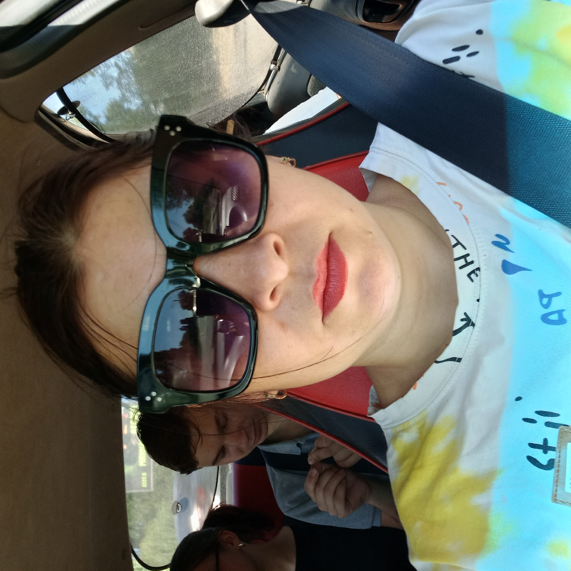 A frontal, eye-level, medium close-up shot shows a woman wearing large, dark sunglasses and a white t-shirt with a tie-dye pattern of yellow, blue, and white. Her lips are painted red, and she is sitting in the passenger seat of a car, with her seatbelt fastened. To her left, partially visible, is another person, also wearing glasses. In the back seat behind her, a young male with short, dark hair is looking down, with his hands clasped together. The interior of the car is visible, including the red and black upholstery of the seats and the car's ceiling. Sunlight streams in from the right side of the vehicle, casting shadows. The scene appears to be outdoors in Kropyvnytskyi, Ukraine, indicated by the reflection in the woman's sunglasses which shows trees and a road, suggesting a daytime journey.