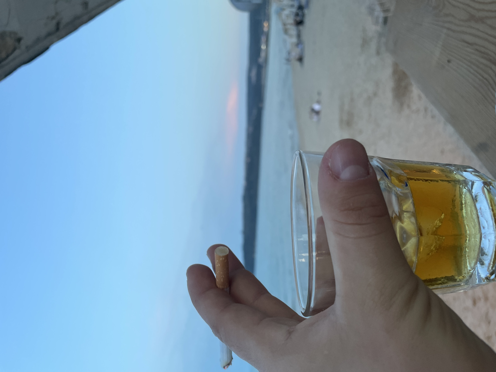 The image presents a first-person perspective from a beachfront setting in Varna, Bulgaria, during late afternoon or early evening. In the foreground, a left hand holds a lit cigarette between the index and middle fingers, its tip glowing red. Simultaneously, the hand grips a clear, faceted glass containing an amber-colored liquid, likely an alcoholic beverage, with visible ice or aeration. The midground features a sandy beach with several indistinct figures, possibly people relaxing on sun loungers or walking, and some white structures. Beyond the beach, the calm, blue-grey water of the Black Sea extends towards a distant landmass or coastline. The horizon reveals a faint pink and lavender hue in the sky, transitioning to clear light blue overhead. Distant urban structures, including a distinctive curved building and some artificial lights, are visible on the far shore. A wooden surface is present in the lower right, and an overhead structure partially intrudes into the upper right corner of the frame.