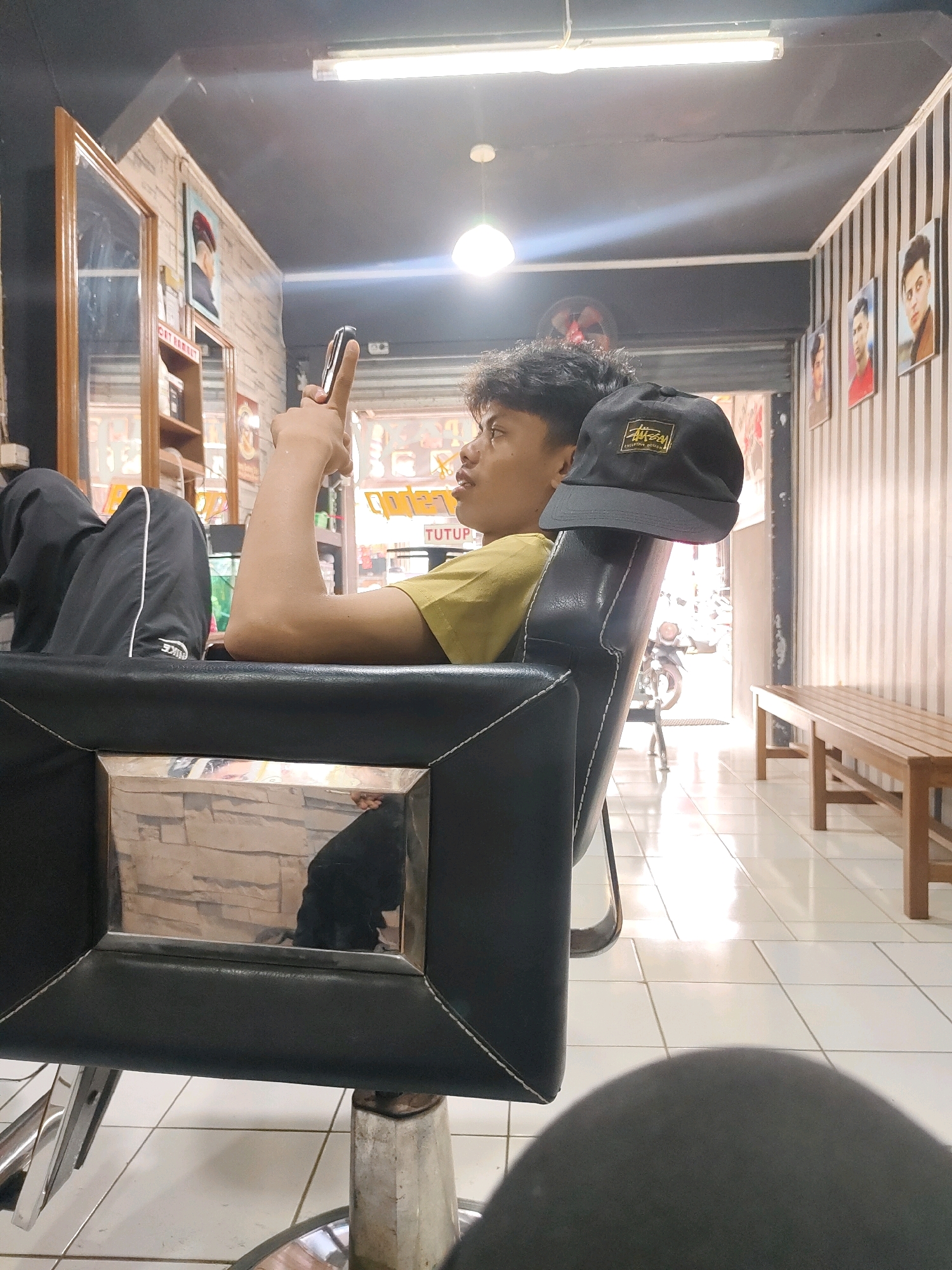 A young male is seated in a barber chair, looking at his phone. He is wearing a yellow t-shirt and black pants. A black baseball cap with a logo is placed on the headrest of the chair. The barber shop is decorated with framed pictures of men on the wall. There is a wooden bench and a motorcycle visible in the background. The floor is tiled. The scene is lit by fluorescent lights and natural light coming from the open doorway. The barber shop is located in Sukanagara, Indonesia.