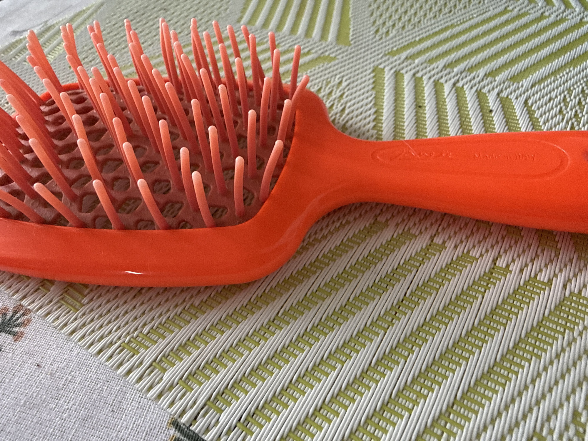 This is a close-up, eye-level, medium shot of an orange hairbrush lying on a textured, woven placemat. The placemat has a repeating pattern of diagonal green and white stripes. The hairbrush is positioned with its handle extending from the right side of the frame towards the left, and the bristled head occupying the left and central portions of the image. The brush has a hollow, open design with many thin, orange bristles. A small, dark speck is visible on the placemat near the bottom left corner of the image. On the handle of the hairbrush, faint text is embossed, reading "Made in Italy". The lighting appears to be natural and diffused, suggesting an indoor setting. There are no people or any other objects visible in the frame. The image focuses solely on the hairbrush and its immediate surroundings.