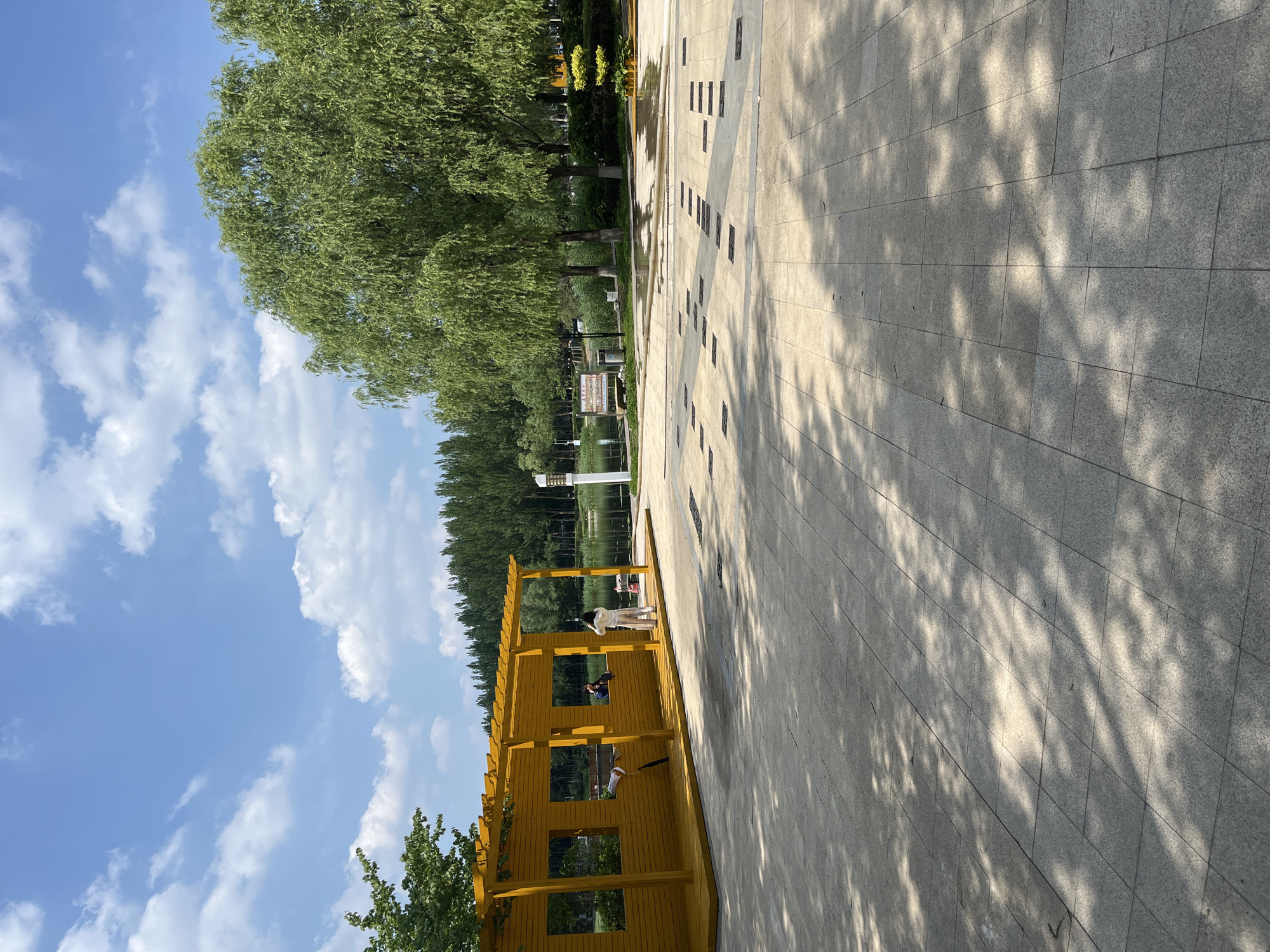 In Changchun City, China, on a sunny day with scattered clouds, a bright yellow, open-sided wooden structure stands on a paved plaza. The structure has a sloped roof supported by posts and features rectangular openings resembling windows. Two individuals are visible within or near this structure: one person, seen from the side, is standing and appears to be taking a photo, while another, partially obscured, is seated inside.
Beyond the plaza, a body of water, possibly a lake or river, is visible, bordered by a line of tall, slender trees in the background. To the right of the water, a weeping willow tree with lush green foliage dominates the scene. The plaza itself is paved with large, light-colored square tiles, with patterns of darker squares and rectangular elements embedded within the paving. Shadows from overhead foliage are cast across the plaza, indicating the time of day. A white lamppost stands near the water's edge, and a wooden bench is situated in the mid-ground. The overall impression is of a park or recreational area on a pleasant day.