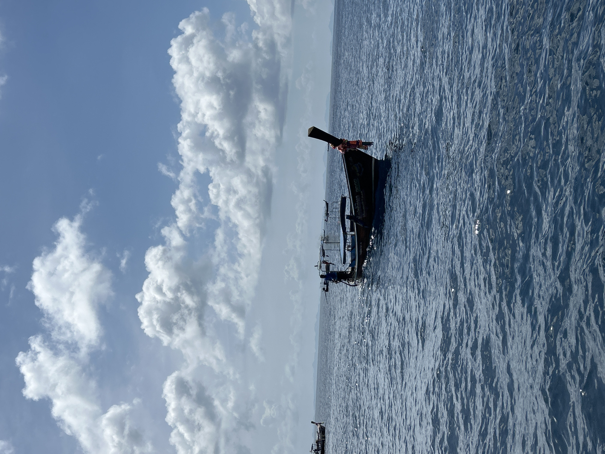 The image depicts a body of deep blue water with a moderate ripple. In the foreground, a traditional Thai longtail boat with a dark hull is prominently featured. A person, wearing a blue shirt and dark pants, stands near the stern of this boat, appearing to operate it. Another longtail boat is visible in the mid-ground to the left, further out on the water. The horizon line is marked by a faint, distant landmass, possibly islands, under a sky filled with scattered white cumulus clouds against a pale blue background. The scene is likely located in the waters surrounding Haad Yao, Thailand, a popular tourist destination known for its beaches and boat tours.