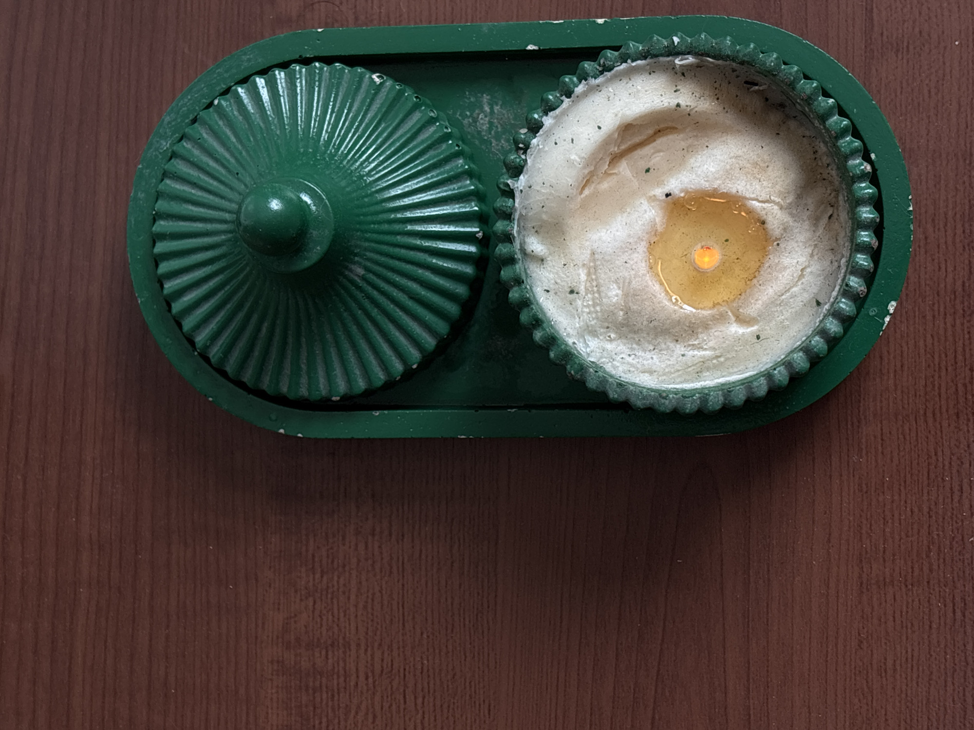 The image depicts a green, divided serving dish on a wooden surface. The dish has two compartments: one is covered with a ridged lid, and the other contains a creamy, textured substance with a clear, yellow liquid pool in the center. The scene is indoors, but specific location information for Santa Clara Ocoyucan, Mexico is not present in the image.