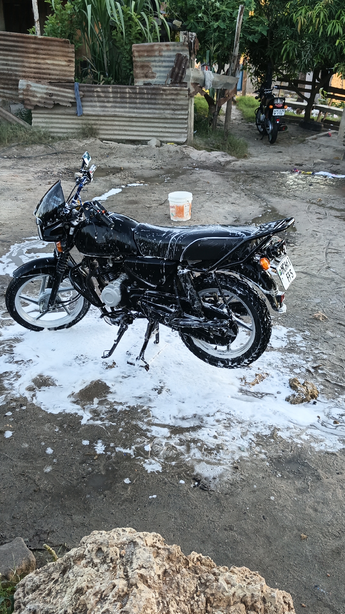 A black motorcycle is covered in white soap suds, likely during a wash, and parked on a dirt surface. A white bucket with orange accents is placed near the motorcycle's seat. In the background, there is a ramshackle structure of corrugated metal and some green vegetation. Further back, another motorcycle is parked near a wooden structure. The license plate of the main motorcycle indicates it is registered in Tanzania. The location may be a residential area in Dar es Salaam, Tanzania.