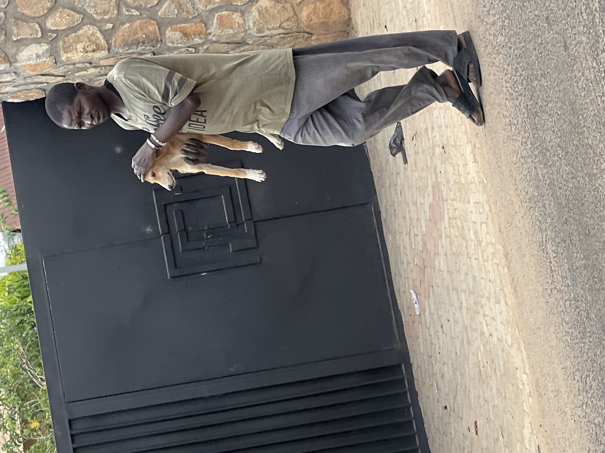 This image, rotated 90 degrees counter-clockwise, depicts a man holding a dog outdoors in Gura Top, Nigeria, during daylight hours on what appears to be a clear day.
The primary subject is a dark-skinned man with closely cropped dark hair, standing and gently holding a light brown and white dog. He is wearing a light olive-green short-sleeved t-shirt that has "IDEA" clearly visible on the front, and the word "Coffee" partially visible above it. He also wears two white bracelets on his right wrist. His trousers are dark grey, and he has black sandals or open-toed shoes on his feet. The dog he is holding has predominantly light brown fur with white markings, particularly on its paws. Its face is light brown with darker features around the eyes and nose.
Behind the man and dog is a tall, dark grey or black metal gate, which appears to be the entrance to a property. The gate has horizontal slats at the bottom and a raised rectangular design in the upper section. Above the gate, a wall constructed from light brown and beige rough-cut stones is visible. The ground where the man is standing is paved with light-colored, possibly beige, square tiles, which transition into a coarser, light-colored concrete or asphalt surface further to the right. Some green foliage is visible on the far left edge of the frame, behind the gate. The man's expression is neutral, seemingly focused on holding the dog.