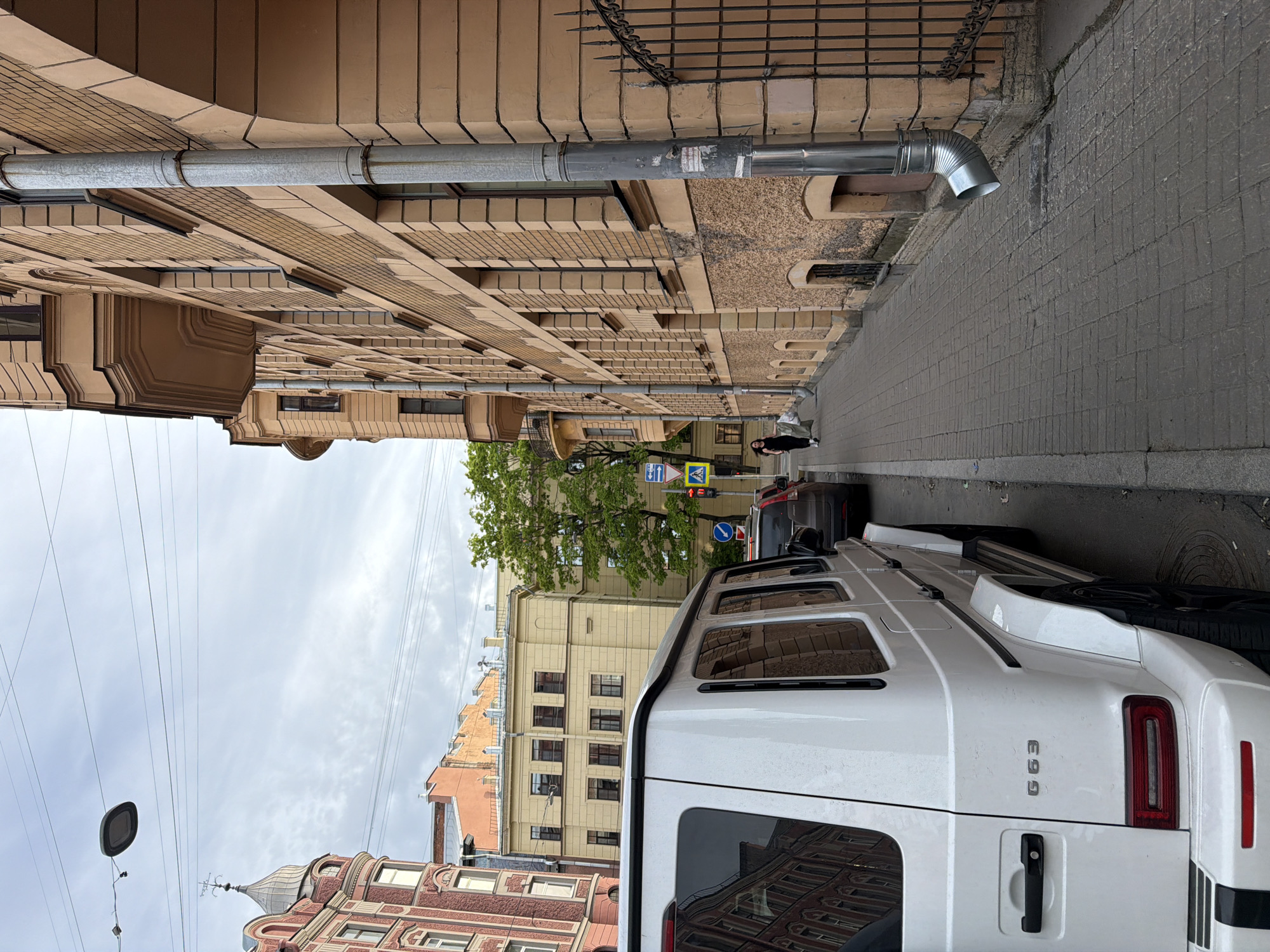 The image captures a narrow city street in Saint Petersburg, Russia, under an overcast sky. In the immediate foreground, a white Mercedes-Benz G63 SUV is parked on the right side of the road, its rear quarter and part of the roof visible. The "G 63" emblem is clearly legible on the rear pillar.
On the left side of the street, a multi-story building with light beige, ornate facades extends upwards. It features numerous windows, decorative architectural elements, and a prominent metallic downspout running vertically along its exterior. Some faint, illegible text or stickers are visible on the downspout.
Further down the street, on the right, another building with a reddish-brown base and a lighter upper section is visible, characterized by a distinctive domed turret topped with an ornate spire. A pedestrian in dark clothing can be seen walking on the sidewalk near the corner. Several road signs are visible: a blue rectangular sign with a white arrow pointing right (indicating a mandatory direction or turn) and a yellow triangular sign with a black pedestrian crossing symbol.
Parked further along the street are additional vehicles, including a dark SUV and another dark car. A small tree with green foliage adds a touch of nature to the urban setting. Overhead power lines crisscross the light grey, cloudy sky. The street itself is paved, with a brick-patterned sidewalk on the right. The overall architectural style and visible road signs are consistent with the location context of Saint Petersburg. The scene depicts a quiet daytime street, with no specific activity taking place beyond general urban life.