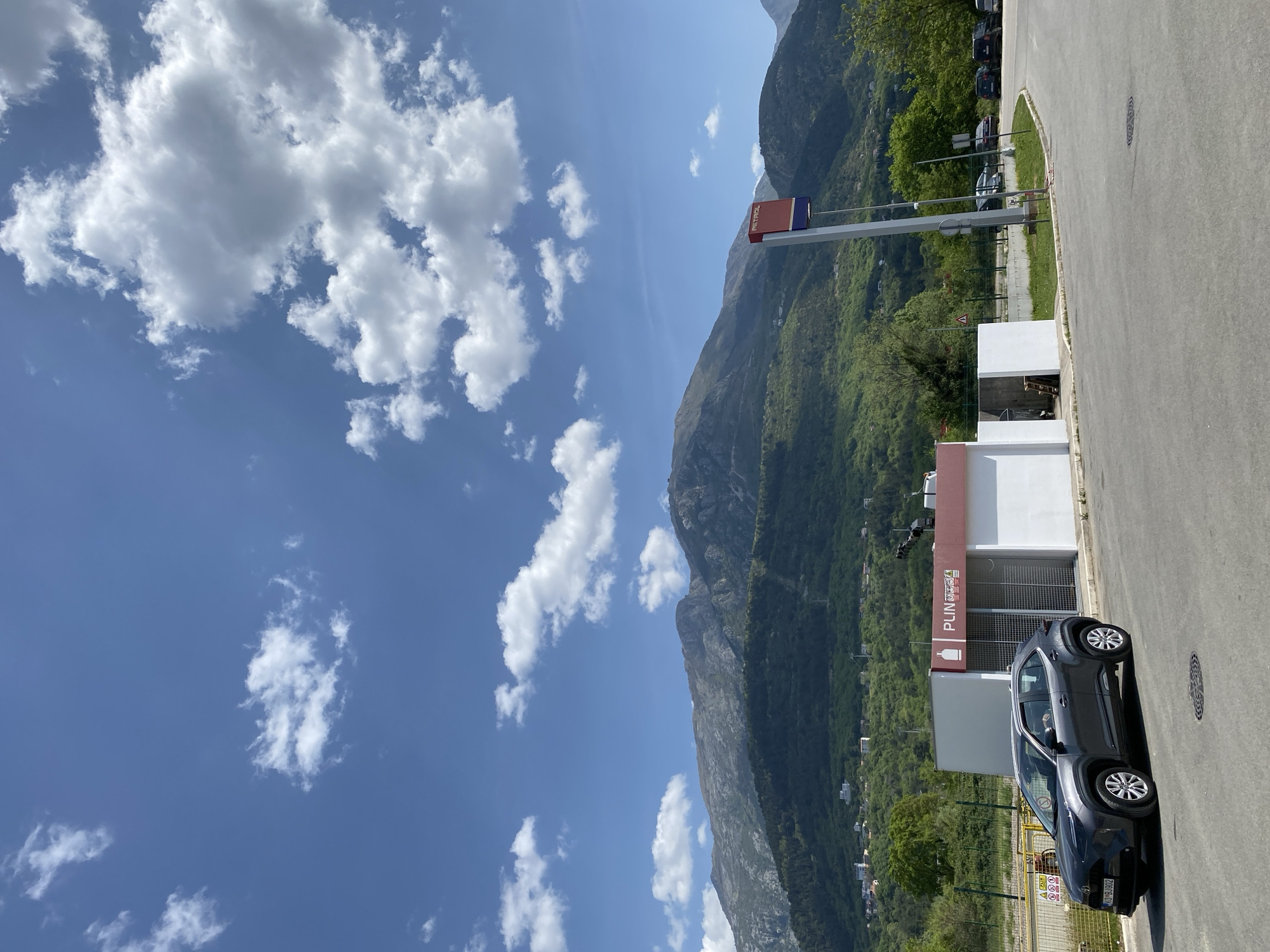 The image displays a sunny daytime scene in a mountainous region, likely Kavač, Montenegro. In the foreground, a dark gray SUV, resembling a Toyota C-HR, is parked on an asphalt lot. Its license plate, "KVB B...902", is partially visible, indicating a Montenegrin registration. To the right of the car is a modern, white building with a red and gray facade. On the building, a sign displays "PUNJENJE" with an icon of a bottle/cylinder, and "AUTOGAS" below it, suggesting it's an LPG or gas filling station. Near the parked car, a yellow warning sign is visible with symbols for caution and fire hazard, and the Cyrillic text "A ЗАБРАНЬЕНО" (Forbidden).
Further back, a tall signpost features a red section with the word "PETROL" and a blue section above it. The background is dominated by large, lush green mountains, characteristic of the Montenegrin landscape. The sky is bright blue with scattered white cumulus clouds. The lighting suggests it is late morning or early afternoon. The scene appears calm and clear, with no people visible. The asphalt area extends into a road or parking lot with green verges and other parked vehicles partially visible in the distance among the trees.