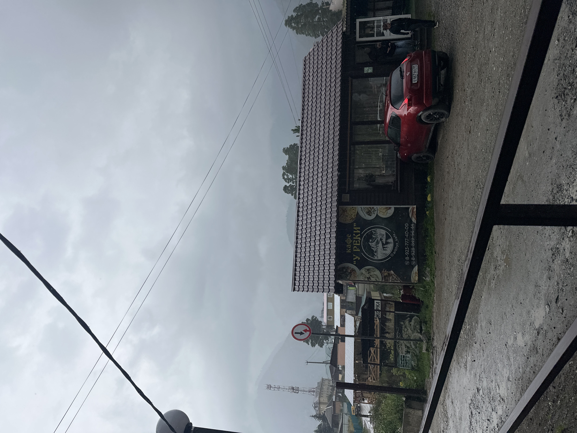 The image shows a street scene in Arkhyz, Russia, on a cloudy day, likely during or after rain as the ground is wet and there are visible raindrops on the cafe's window. In the foreground, a dark metal railing runs horizontally across the bottom of the frame. Behind the railing is a gravelly, wet road with puddles. To the right, a small, red Nissan Juke is parked on the gravel. Three people are visible near the red car and by the entrance of a small building.
The building in the midground is a single-story structure with a dark brown, tiled roof. It appears to be a cafe, as indicated by signage. A prominent sign on the left side of the building displays the text "КАФЕ 'У РЕКИ'" (Cafe "By the River") in Cyrillic, along with a circular logo featuring a mountain landscape. Below the cafe name, phone numbers are listed: "8-923-777-40-00" and "8-928-849-94-44". A social media handle "@arkhyz_kafe" is also visible on the sign.
Further in the background, several other buildings, including a multi-story structure and smaller wooden houses, are visible. A tall communication tower stands prominently on a distant, mist-covered mountain. Trees are scattered throughout the scene, and a road can be seen curving to the left, with a yellow vehicle partially visible. A traffic sign with red and white circles and arrows indicating lane direction is positioned near a tree. Overhead, power lines stretch across the frame against the overcast sky.