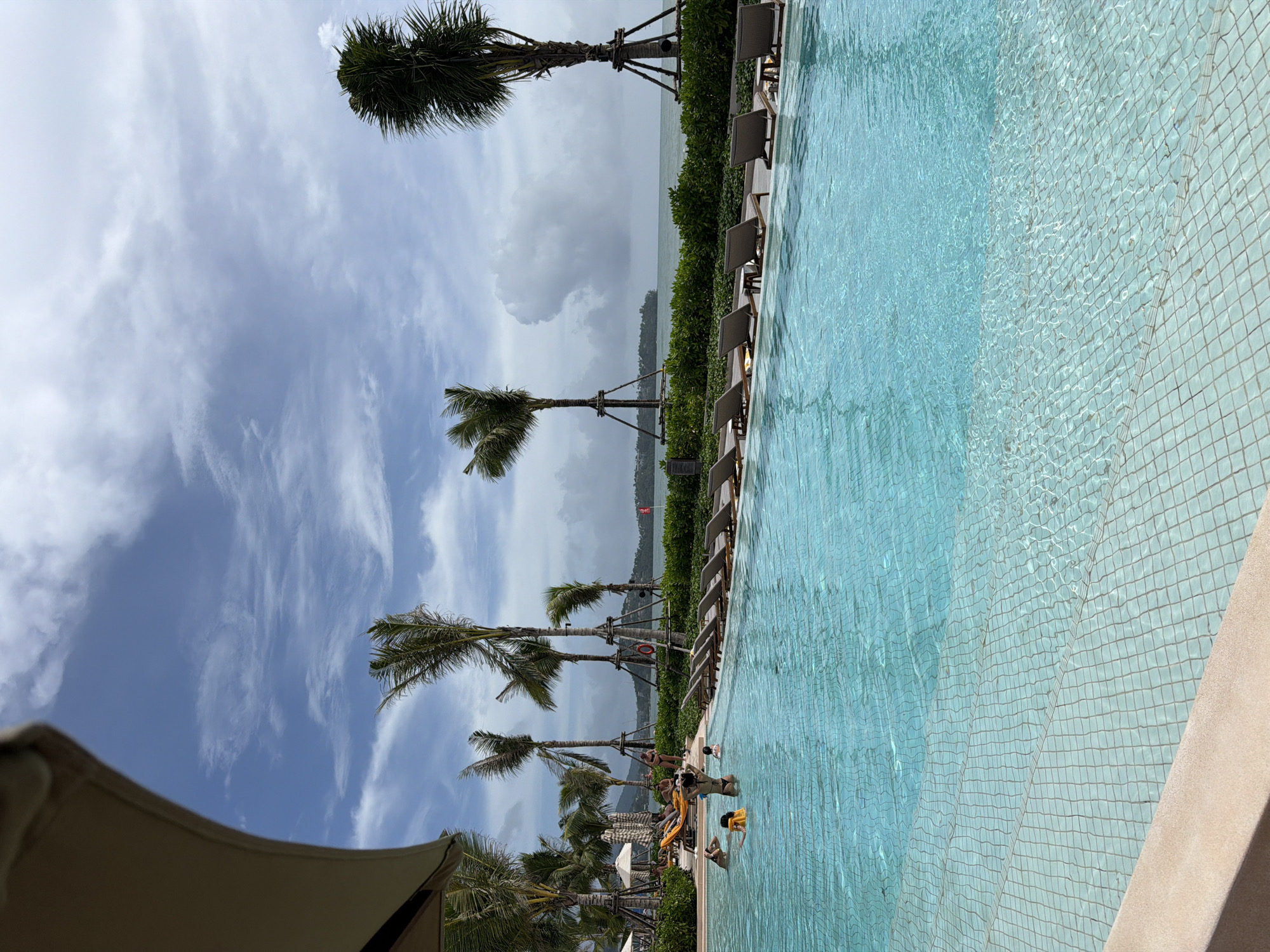 The image depicts a swimming pool in Choeng Thale, Thailand. The pool is tiled with small, light blue squares, and the water is clear. Adjacent to the pool are lounge chairs positioned next to a line of green bushes and palm trees. In the background, there is a view of a body of water under a cloudy sky. Several people are in the pool.