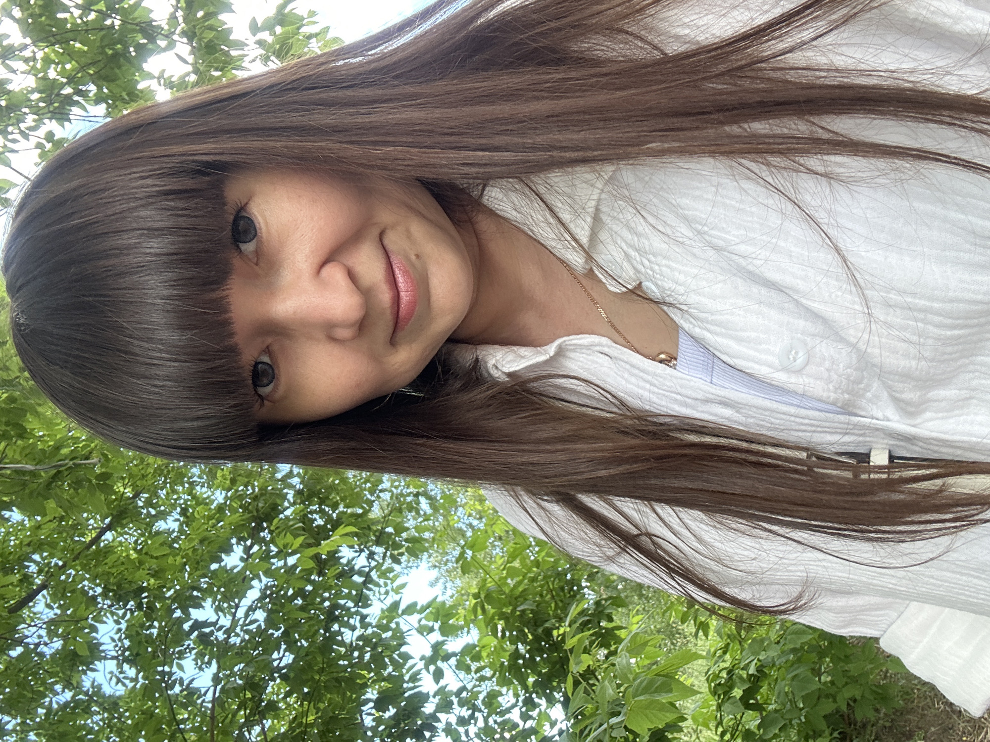 A direct portrait shot captures a female subject outdoors in the city of Chaikovsky, Russia. She possesses long, straight, dark brown hair with blunt bangs and appears to have grey contact lenses in her eyes. Her expression is a subtle smile. She is dressed in a white, textured, long-sleeved collared shirt, unbuttoned to reveal a lighter garment underneath, and wears a gold-colored chain necklace with a small, indistinct pendant. The background features abundant green foliage from trees, with visible patches of clear blue sky above. The scene is illuminated by natural daylight.