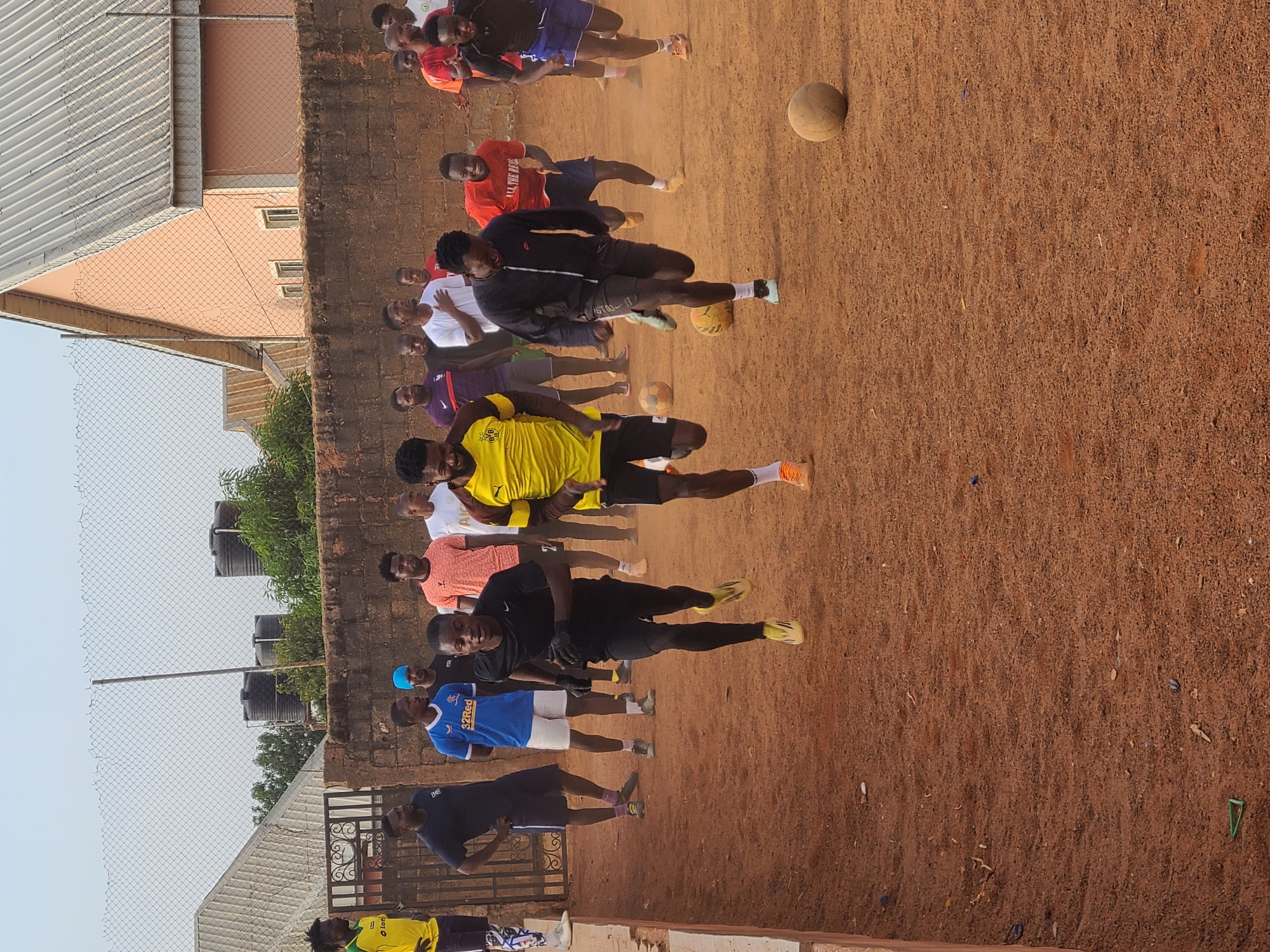 A medium-shot, eye-level captures a group of approximately 15 men practicing soccer on a dirt field in an urban environment. The men are variably dressed in soccer jerseys (yellow, blue, red) and sports attire (shorts, t-shirts).
Several soccer balls are visible in the foreground, scattered among the men as they run and kick. A brick wall stands parallel to the background, with water tanks and greenery rising above it.
A metal fence is on the left, and the roofs of houses/buildings rise above the fence, indicating an urban setting. The sky is overcast.
The location is Unknown, Unknown.