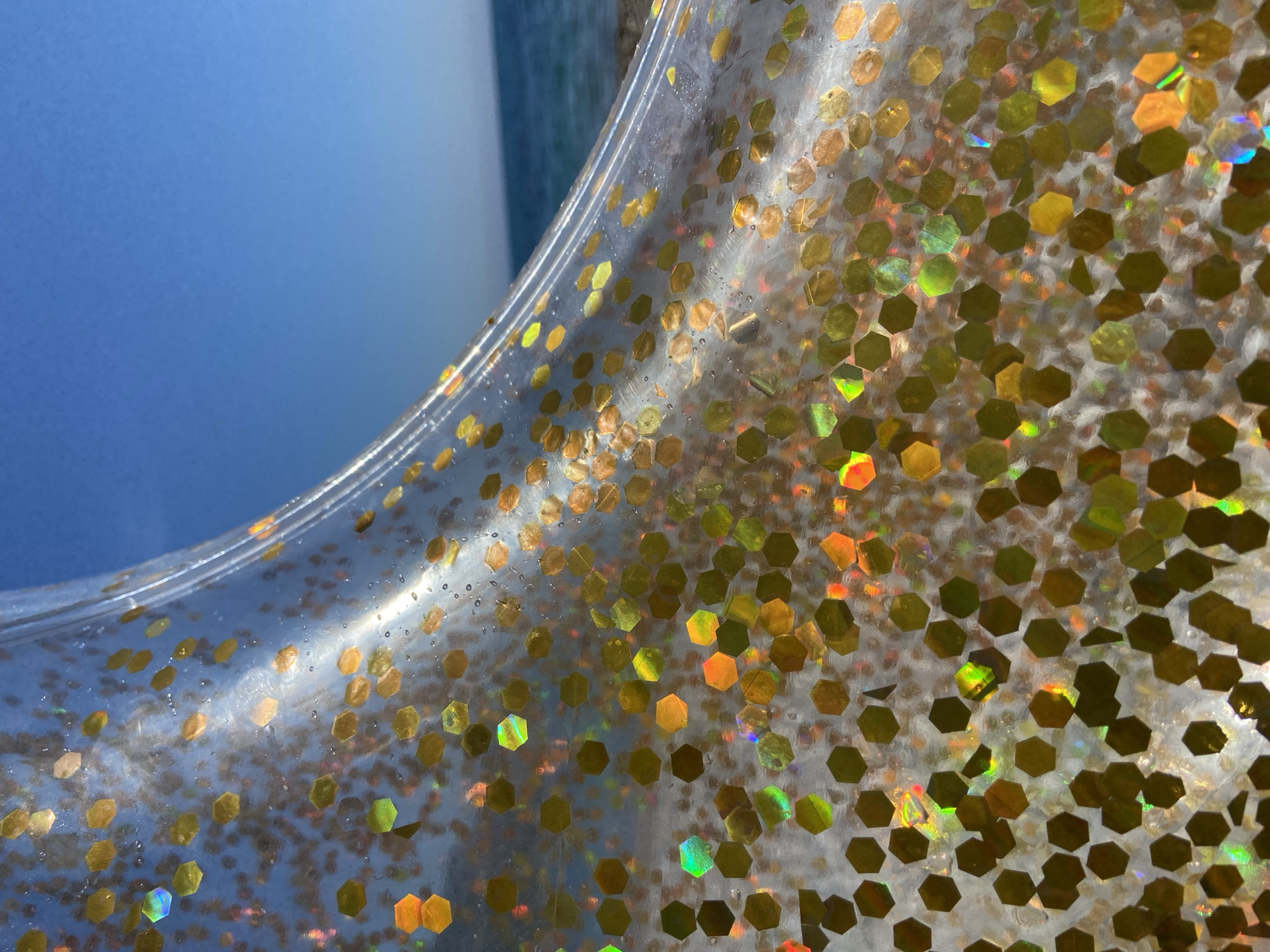 The image presents a close-up view of a clear, translucent material densely packed with numerous hexagonal glitter pieces. The glitter is predominantly golden or yellow, but reflects an array of iridescent colors including green, orange, red, and blue, indicating strong interaction with light. These hexagonal pieces vary slightly in size and are suspended within the clear material, which shows subtle curves, suggesting it is flexible. Small air bubbles or imperfections are also visible within the transparent substance.
In the background, a large, blurry expanse of bright blue is visible, likely representing a clear sky or body of water, suggesting an outdoor setting under bright daylight conditions. A less defined, lighter area is also visible towards the left side of the frame in some views. There is no specific activity or event taking place, and no people or visible text are present. The scene is static, highlighting the texture and reflective qualities of the glitter-filled material. The location context provided is Prijevor, Montenegro.
