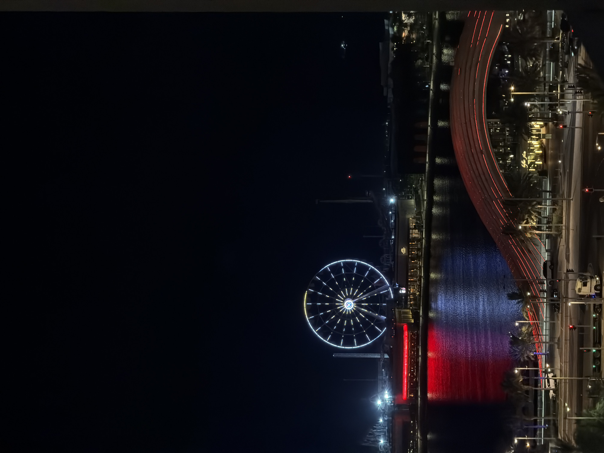 This is a night time, medium shot, eye-level, vertical, exterior photograph of a Ferris wheel and a waterfront scene. The Ferris wheel is illuminated with white and yellow lights and is the most prominent object in the photograph, positioned in the upper center. The Ferris wheel is located in an amusement park, with other rides visible in the background. The waterfront is illuminated with red and blue lights, creating reflections on the water. Palm trees and streetlights are visible along the waterfront, with cars and a truck on the road. A bridge with red lights is also visible in the foreground. The sky is dark, and there are no people visible in the photograph.