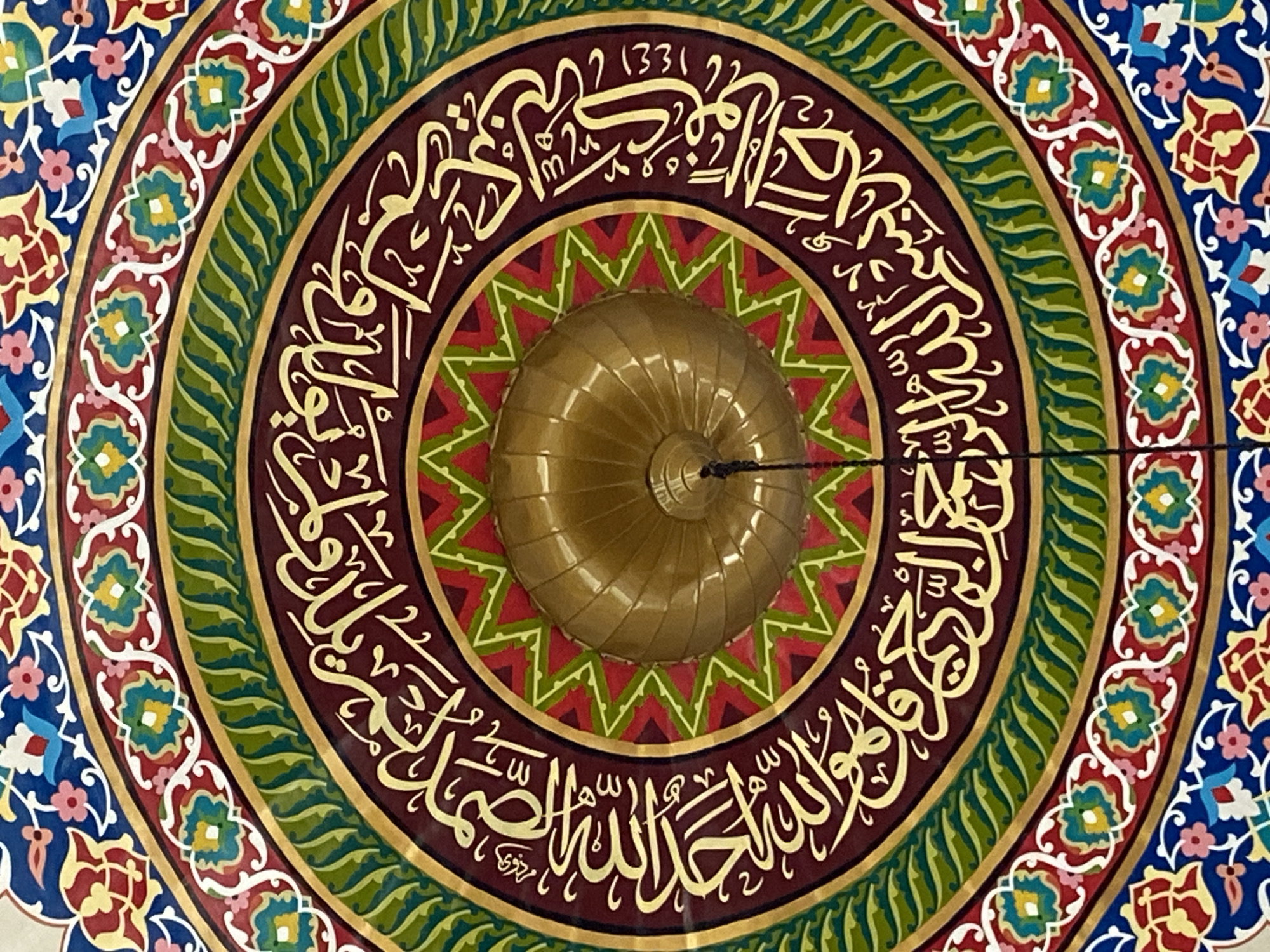 The image displays the interior of a dome, likely within a mosque, featuring intricate Islamic calligraphy and geometric patterns. The calligraphy, rendered in gold on a dark red background, is arranged in a circular band. Surrounding this band are concentric decorative elements: a green wavy pattern, a thin gold border, a white scalloped border with colorful floral motifs in red, green, and blue, and an outer border of blue with red and yellow floral designs. At the center of the dome, a golden, ribbed dome structure is visible, with a black cord hanging down from its apex. The overall impression is one of rich ornamentation and religious artistry. This specific artwork is located at the Baiturrahman Grand Mosque in Lhokseumawe, Indonesia.