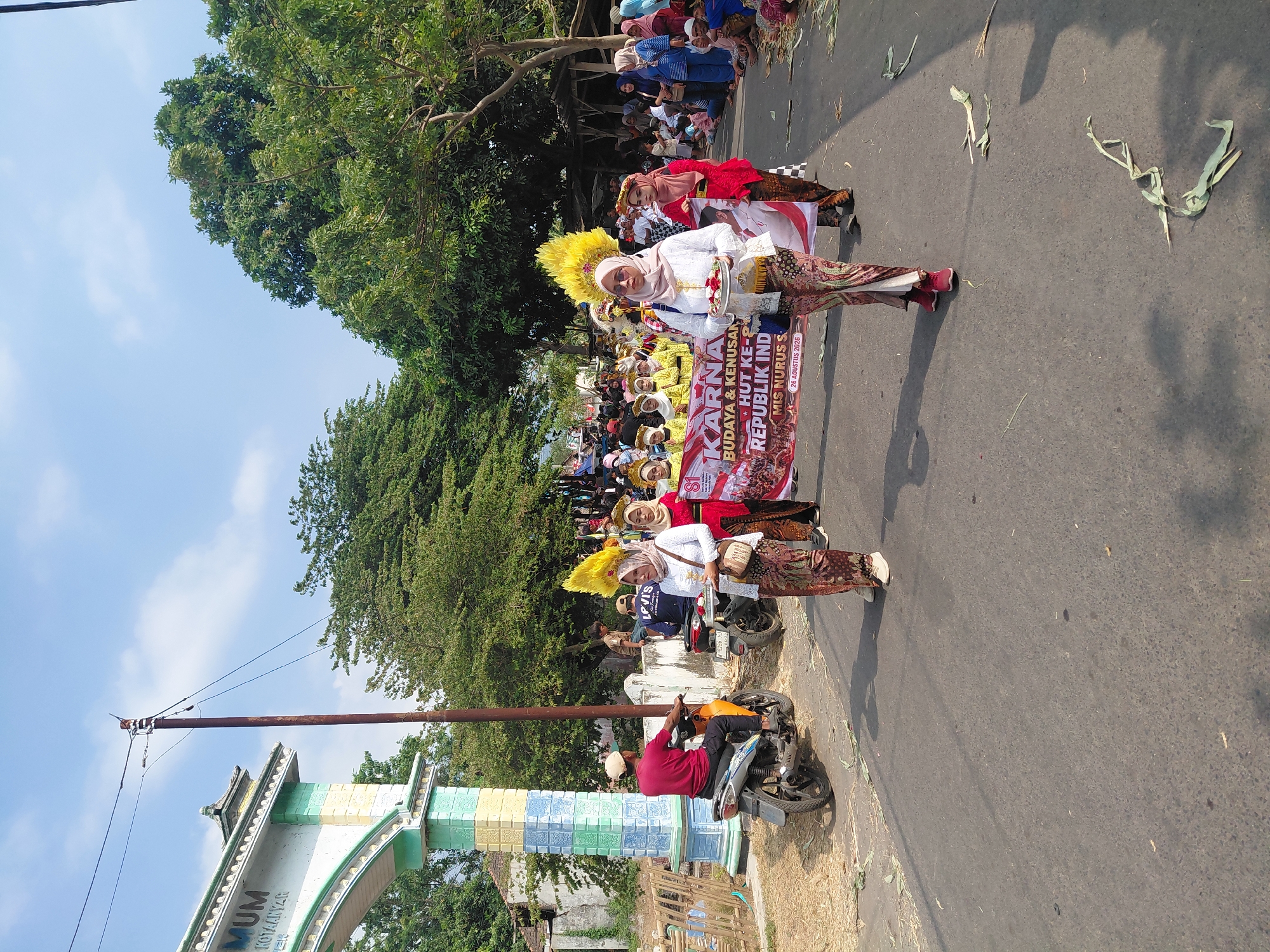 In Bimo, Indonesia, a parade procession is underway along a paved street under a clear, bright blue sky. The scene is dominated by a large, colorful banner held by a group of women walking in unison. The banner, predominantly red with white lettering, proclaims "KARNA BUDAYA & KENUSAN HUT KEPULAUAN REPUBLIK IND" and includes smaller text indicating "MIS NURUS S" and "26 AGUSTUS 2026".
The women are dressed in traditional attire. Several wear white tops and patterned skirts, accessorized with elaborate yellow headdresses. Others are in red tops and patterned skirts, also adorned with headdresses. One woman in the foreground, wearing a white top and a patterned skirt, carries a silver tray with red flowers. Another woman beside her, in a red top and patterned skirt, holds a large white flag.
On the left side of the street, a man on an orange motorcycle, wearing a red shirt and a white cap, is heading away from the camera. Behind him, another motorcycle is visible. Further down the street, a crowd of onlookers lines the sides, some seated and others standing, observing the parade.
The environment includes lush green trees lining the street and an archway structure on the far left, decorated with pastel colors. Scattered on the road are dry plant materials. The overall impression is of a vibrant cultural event.