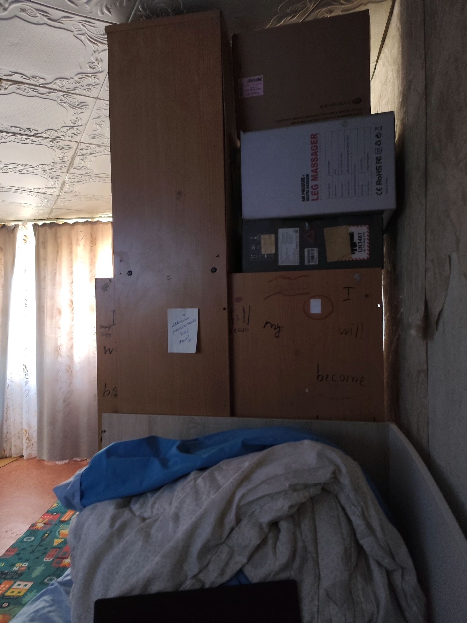 The image shows a cluttered room with stacks of boxes and furniture, including a tall wooden cabinet. A light-colored curtain is visible behind the cabinet. On one of the wooden panels of the cabinet, there are handwritten messages. A crumpled note is attached to the cabinet. In the foreground, a bed is covered with blankets and a blue sheet, with a patterned rug partially visible on the floor. The ceiling appears to be decorated with embossed tiles. The overall impression is of a dimly lit, lived-in space. It is not possible to determine the city of Balqaş, Kazakhstan from the image itself.