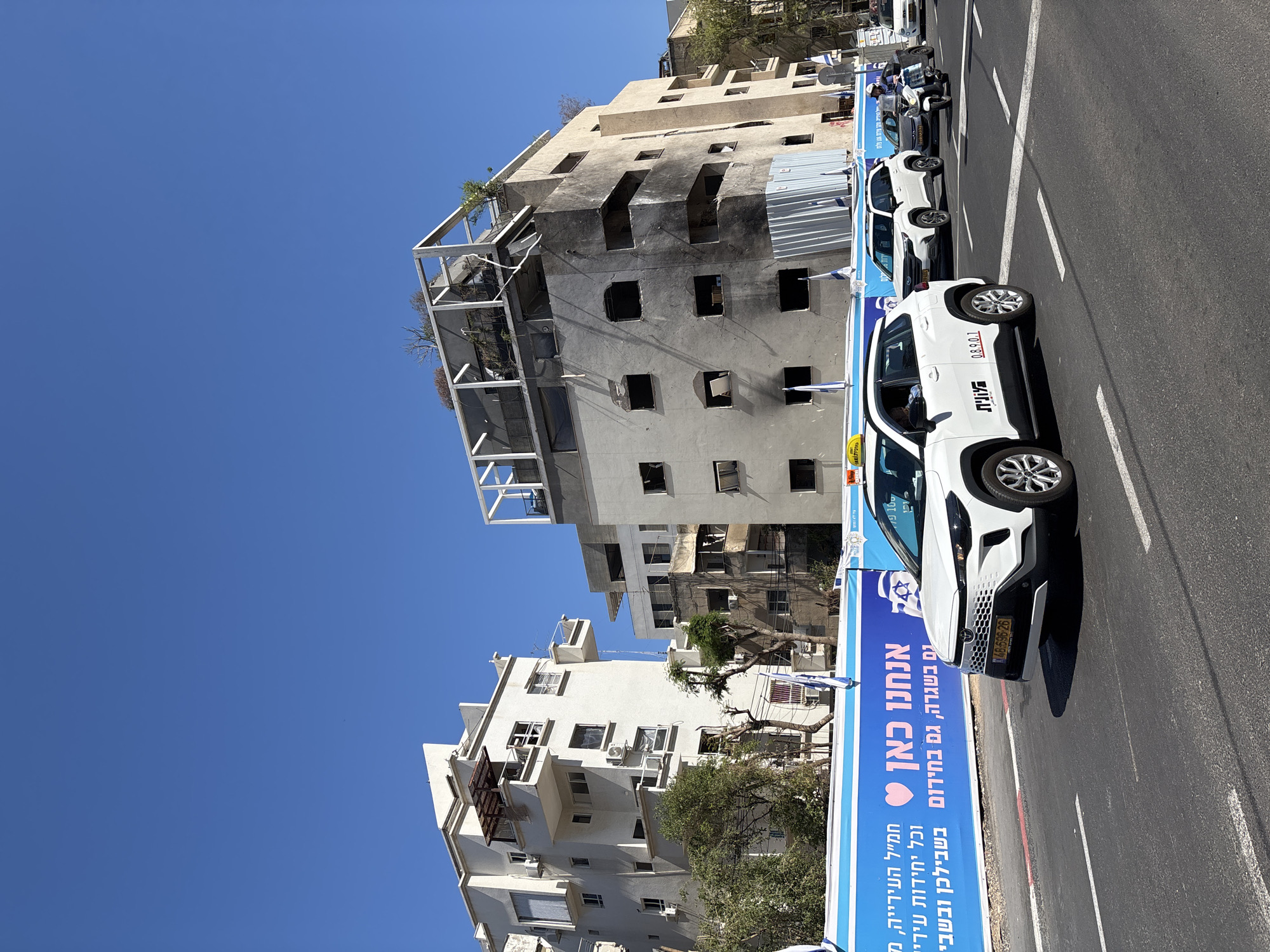 The image captures a street scene in Tel Aviv, Israel, with a clear blue sky overhead. On the right side of the street, several white taxi vehicles are parked in a line, some displaying the Hebrew word "TAXI". Parallel to the parked taxis is a large, blue and white banner with Hebrew text. The banner also contains a Star of David symbol.
In the background, there are two buildings. One is a white residential building with balconies, air conditioning units, and trees around it. Adjacent to the white building is a taller building with a visibly damaged facade, showing signs of fire damage, with blackened areas around the empty window openings. The top of this building has exposed metal framework.
The street itself is asphalt and appears to be in good condition, with lane markings visible. There is no clear indication of human activity in the immediate scene, although the presence of taxis suggests potential transportation activity.