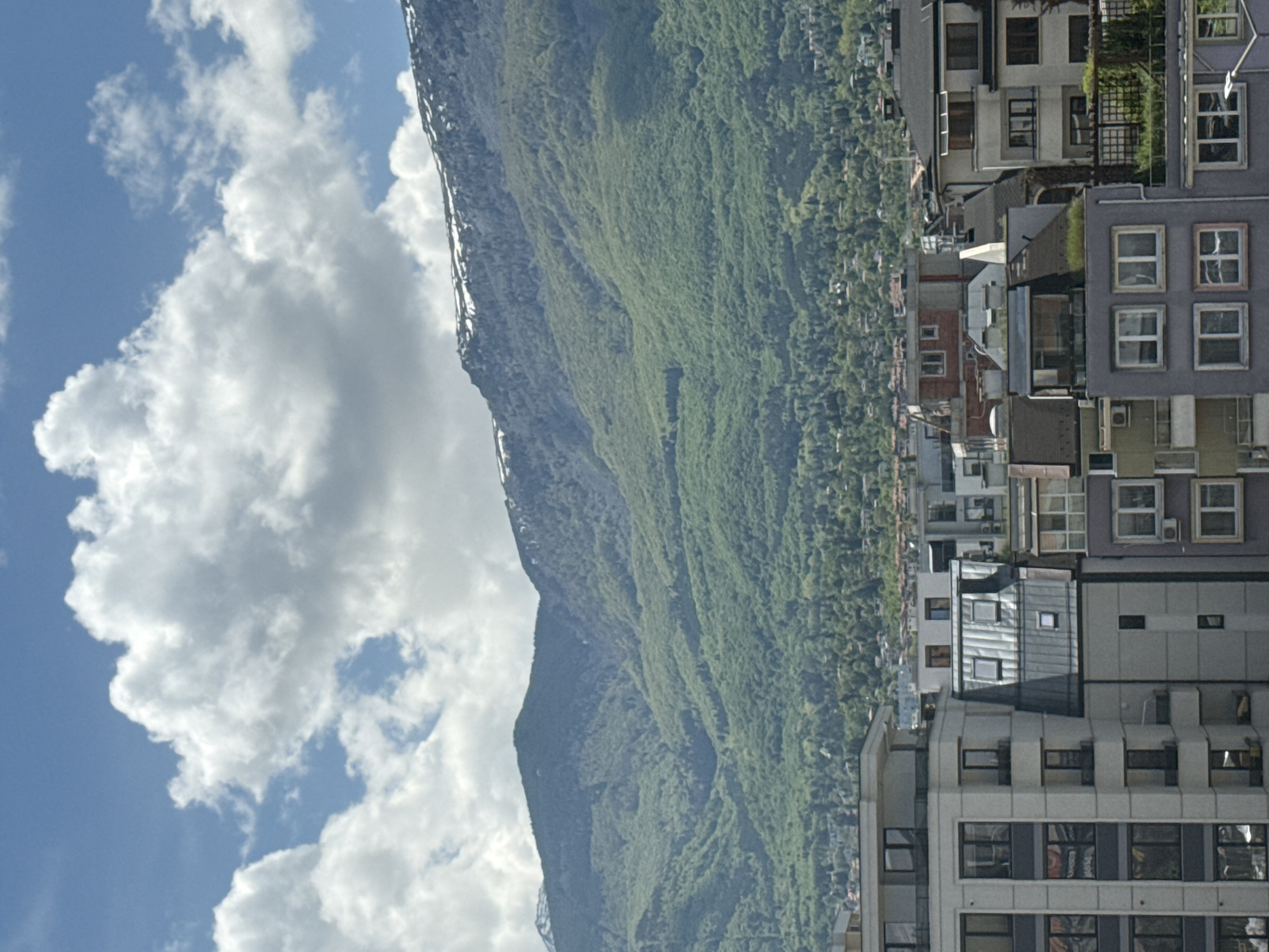 The image presents a view of a city situated at the base of a mountain, with buildings on the right and the mountain ascending to the left. The sky, with a combination of blue and white clouds, takes up a significant portion of the left side of the image. The mountain is covered in green vegetation, and its elevation extends into the sky. Adjacent to the mountain, and further to the right, are densely packed buildings of varied heights and architectural styles. The photograph seems to be taken from an elevated position, overlooking the urban landscape toward the natural terrain. The specific location in the city is not specified, but it is located at the base of a mountain.