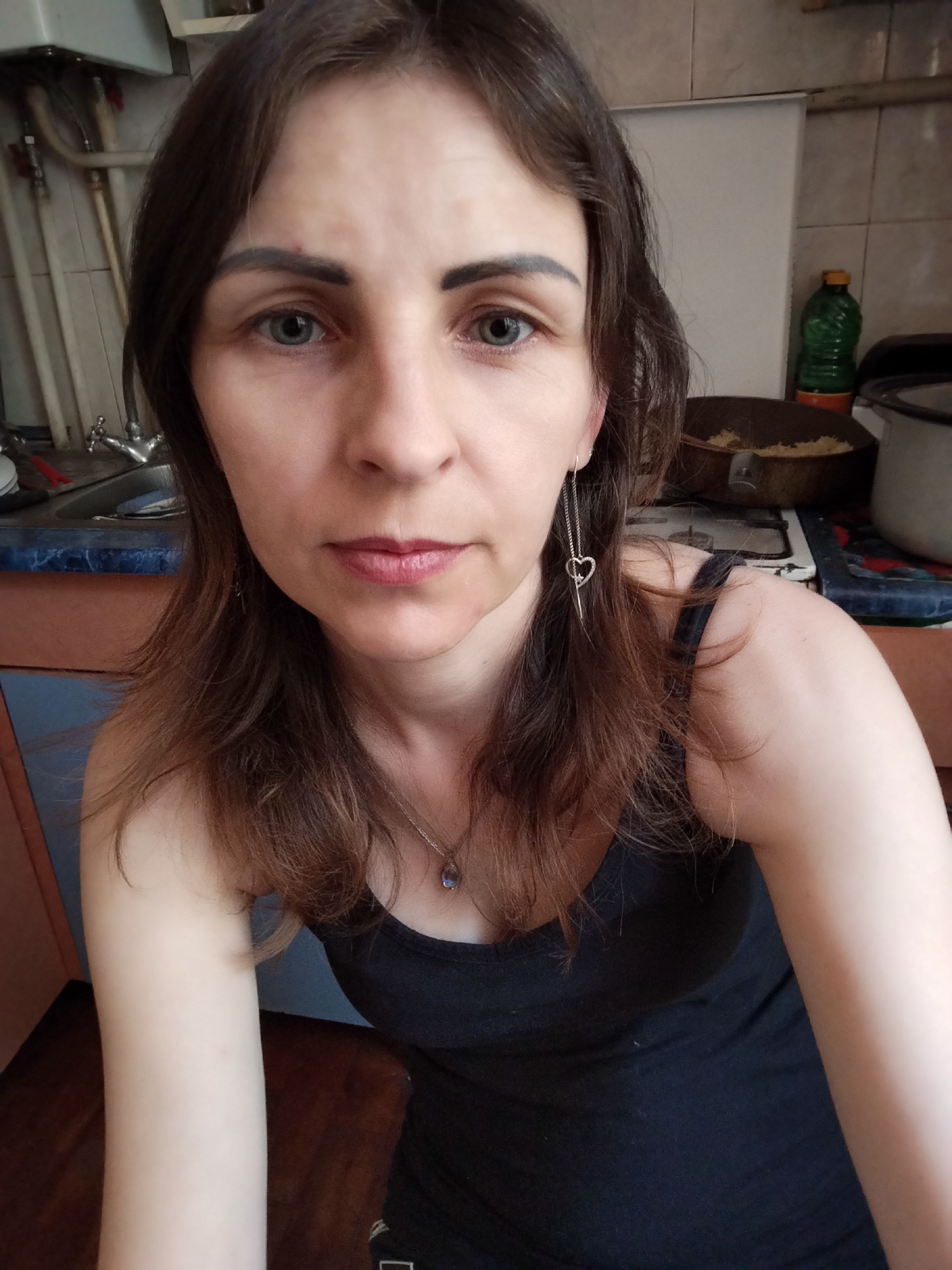 A medium-shot, selfie-style photograph features a woman with dark brown hair, fair skin, and blue eyes. She is wearing a black tank top and a necklace with a dark pendant. A dangling earring with a heart charm is visible on her left ear. Her facial expression is neutral.
The background suggests an indoor setting, possibly a kitchen. To the left, a sink and faucet are partially visible, along with some plumbing. To the right, a stovetop with cookware and a green plastic bottle is present. The overall environment appears somewhat cluttered.
The image is taken from a slightly elevated angle, looking down at the subject. No specific landmarks or identifying features of Vinnytsia, Ukraine, are discernible in the scene.