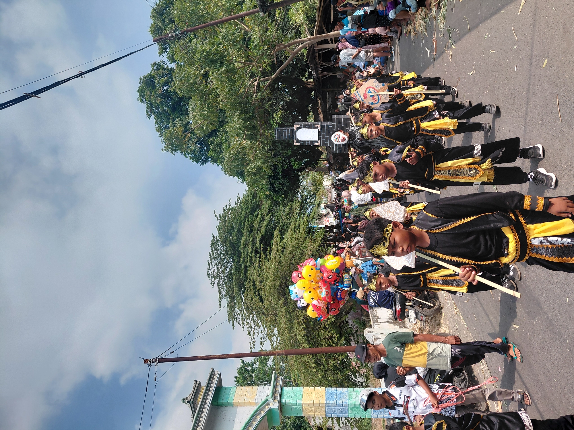 In Bimo, Indonesia, a parade is underway on a paved street under a partly cloudy sky. A group of young boys, dressed in matching black and gold traditional attire with golden headbands, are marching in procession. They are holding wooden staffs and decorated fans. Behind them, a man in a black costume with a white mask and a tall black structure on his back, resembling a tombstone, is also part of the parade. A cluster of colorful balloons, featuring characters like Spider-Man and various cartoon animals, is visible to the left of the procession. Spectators are gathered on the side of the street, some sitting and some standing. The background features trees and a building with a distinctive patterned archway. A motorcycle is parked on the side of the road. The scene captures a lively cultural event with elements of traditional performance and community gathering.