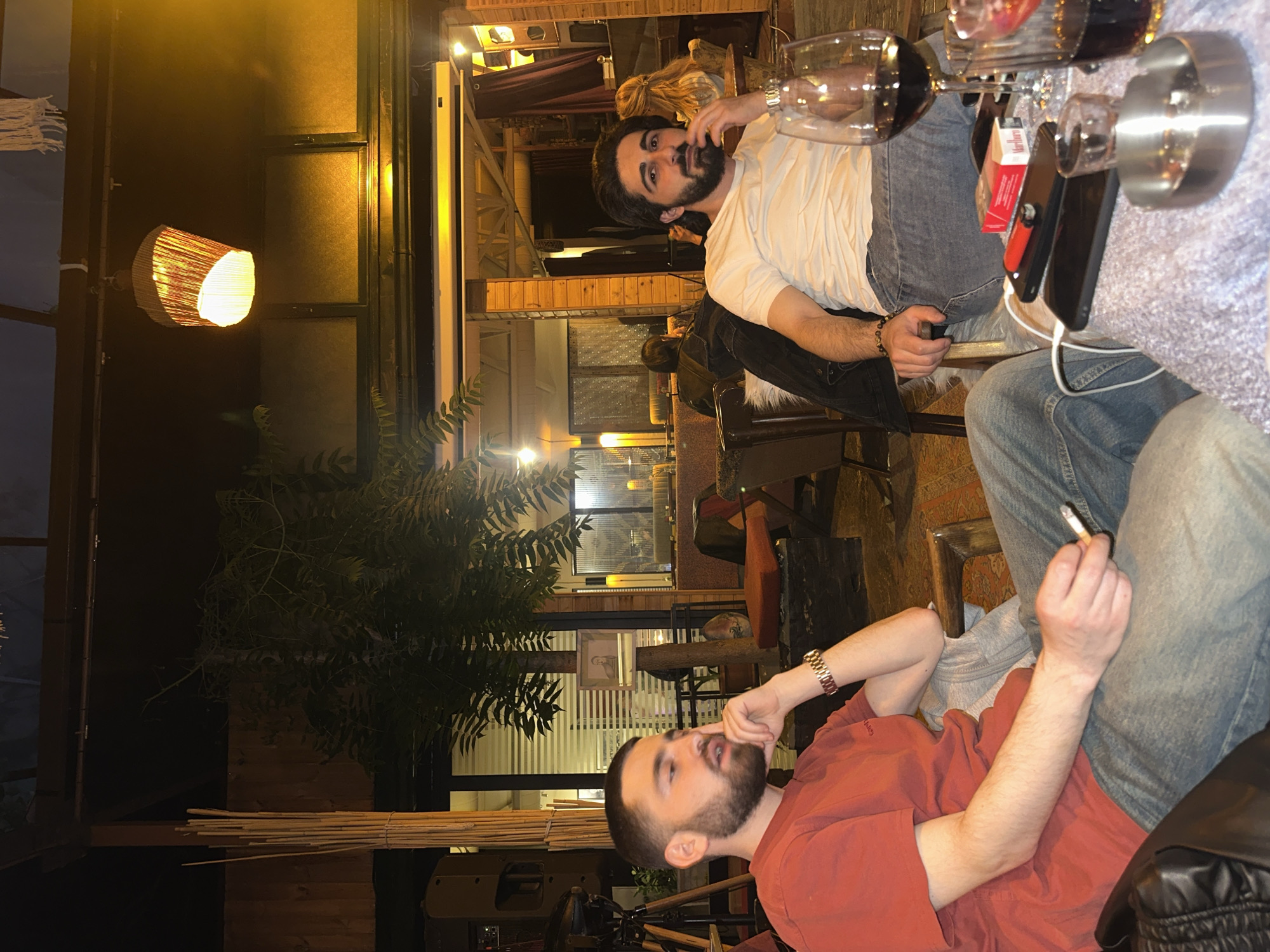 Two men are sitting at a table in an outdoor setting, likely a restaurant or patio. One man, wearing a white t-shirt, jeans, and a black jacket, is seated at the table with two glasses of red wine and a pack of cigarettes next to him. He has dark hair and a beard. The second man, wearing an orange shirt and jeans, is sitting nearby, holding a cigarette and looking thoughtful. A lit lamp hangs nearby. The location is likely in Yerevan, Armenia.
