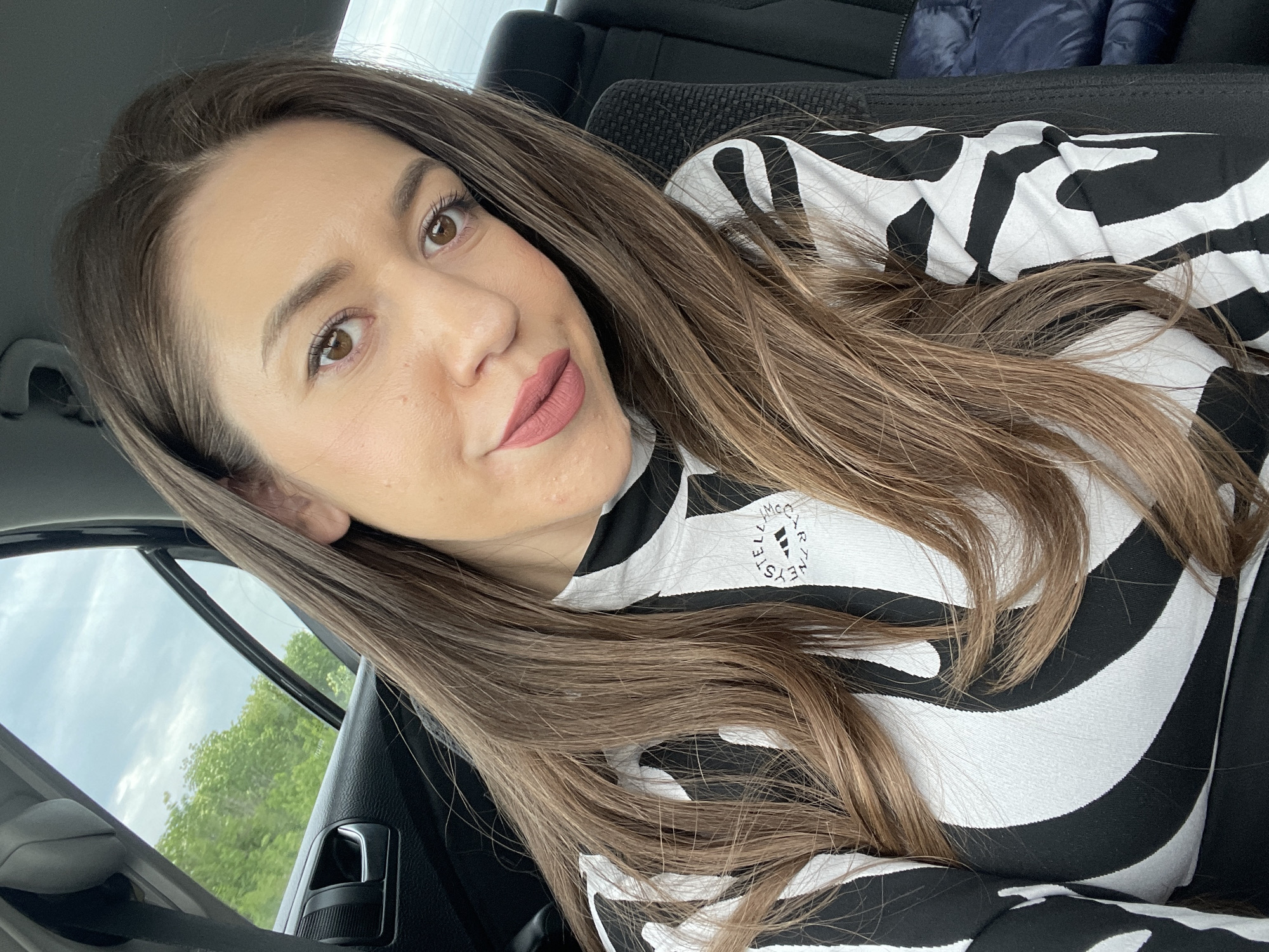 A woman is taking a selfie in the front seat of a car. She has long brown hair and is wearing a black and white zebra print top with an Adidas logo on the upper left. The background visible through the car window suggests a daytime scene with trees and a cloudy sky. The interior of the car is visible, with the dashboard and a portion of the door. There's no information to suggest this image was taken in Yablanitsa, Bulgaria.
