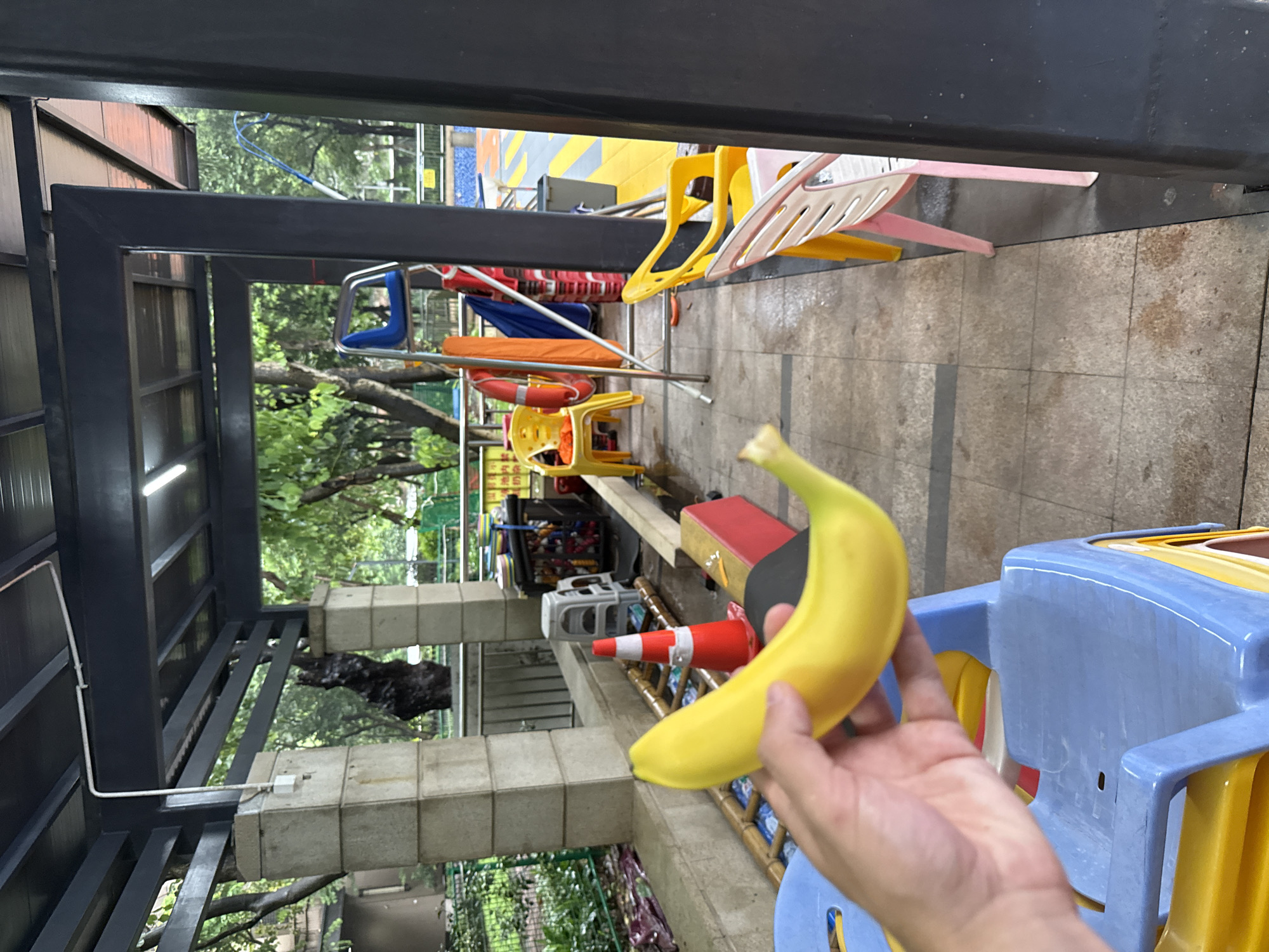 A first-person perspective shot shows a hand holding a yellow banana over a stack of blue and yellow plastic chairs. The scene appears to be outdoors, likely in a public area with a covered walkway. In the background, there are trees, a swimming pool, and various chairs, including yellow and pink plastic ones. The ground is paved with tiles. A lifeguard stand and a red traffic cone are also visible. The overall impression is of a casual, possibly recreational setting. The image appears to have been taken in Haizhu District, China.