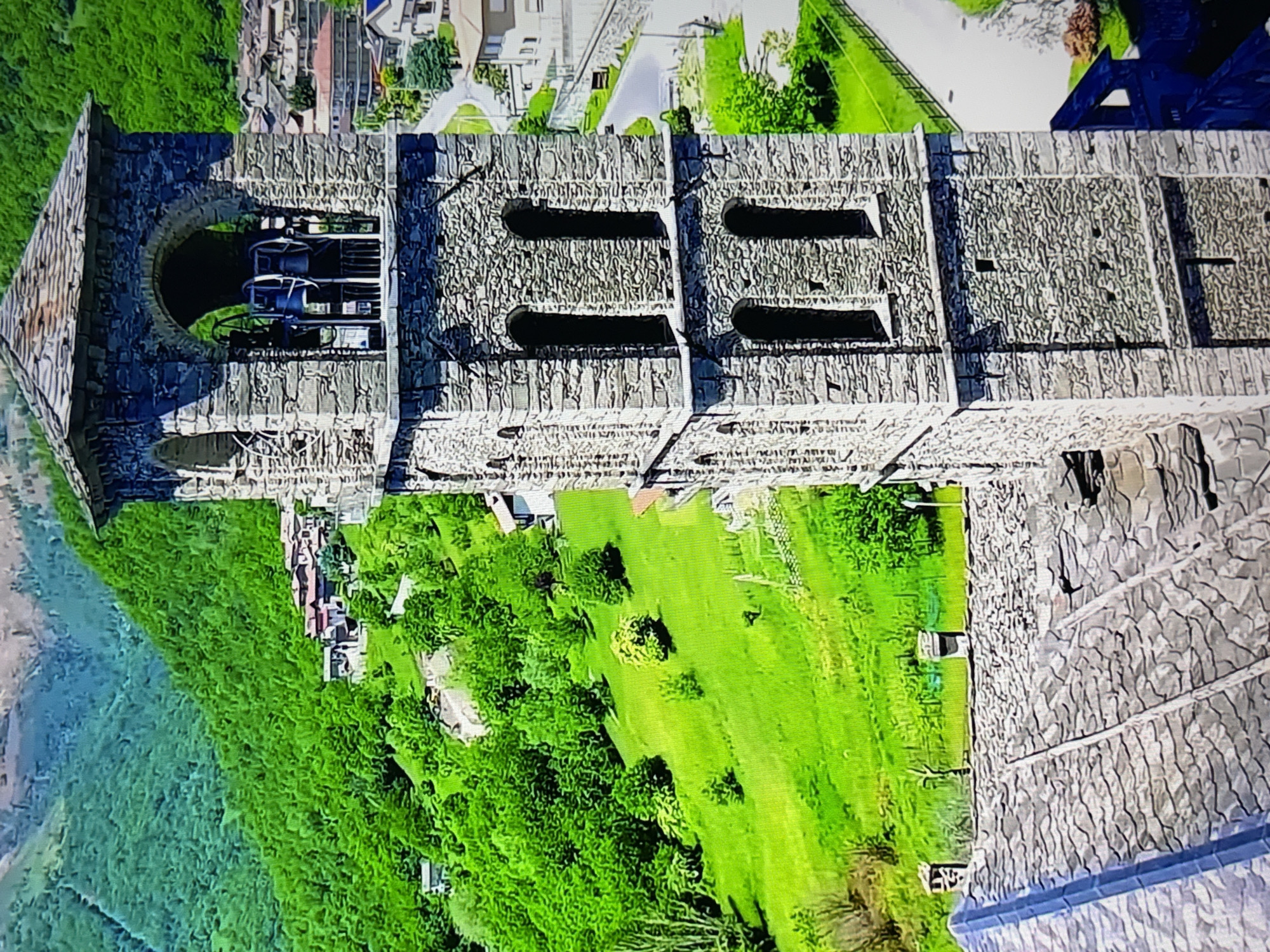 The image captures an aerial view of a stone bell tower, characteristic of a European village. The tower is constructed from rough-hewn stone blocks, featuring arched openings on its upper section, within which bells are visible. Below the arched openings, there are rectangular windows. The tower is capped with a multi-layered stone roof. In the foreground, a section of a building's grey, slate-tiled roof is visible.
The bell tower stands prominently against a backdrop of a lush green, rolling hillside dotted with trees. Small houses with red-tiled roofs are nestled into the slope and further up the hill, forming a village. A winding road is visible in the middle ground, bordered by a fence and grassy areas. The overall environment suggests a rural or semi-rural setting in a mountainous or hilly region, likely in a Mediterranean or Alpine climate, given the stonework architecture and the vibrant greenery. There are no people visible in the image.
The prompt requests information about the location in the city of Burgas, Bulgaria. However, the visual elements of the image do not provide any specific indicators that would place it in Burgas, Bulgaria. The architecture, landscape, and overall style are more commonly associated with other regions of Europe. Therefore, based solely on the visual evidence in the image, it is not possible to confirm its location as Burgas, Bulgaria.