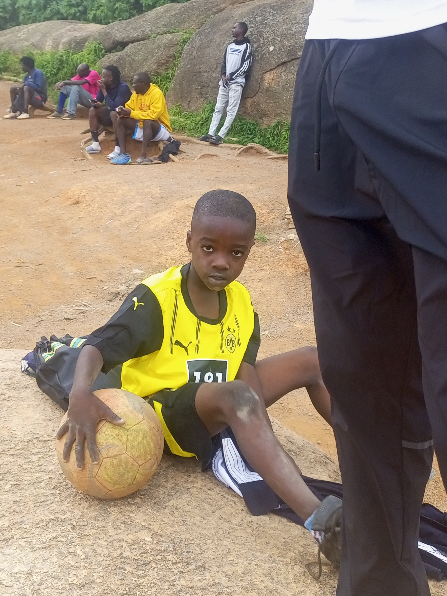 The image depicts a group of young men and boys in Jos, Nigeria, likely gathered for a football practice or event. In the foreground, a young boy wearing a yellow and black football jersey with the number '101' on it sits on the ground, holding a well-worn football. He is positioned next to a black sports bag. Behind him, other individuals are seated on rocks and a wooden structure, some appearing to be resting or observing. The environment is characterized by rocky terrain and sparse vegetation, typical of the landscape in Jos. The overall atmosphere suggests a casual outdoor activity.