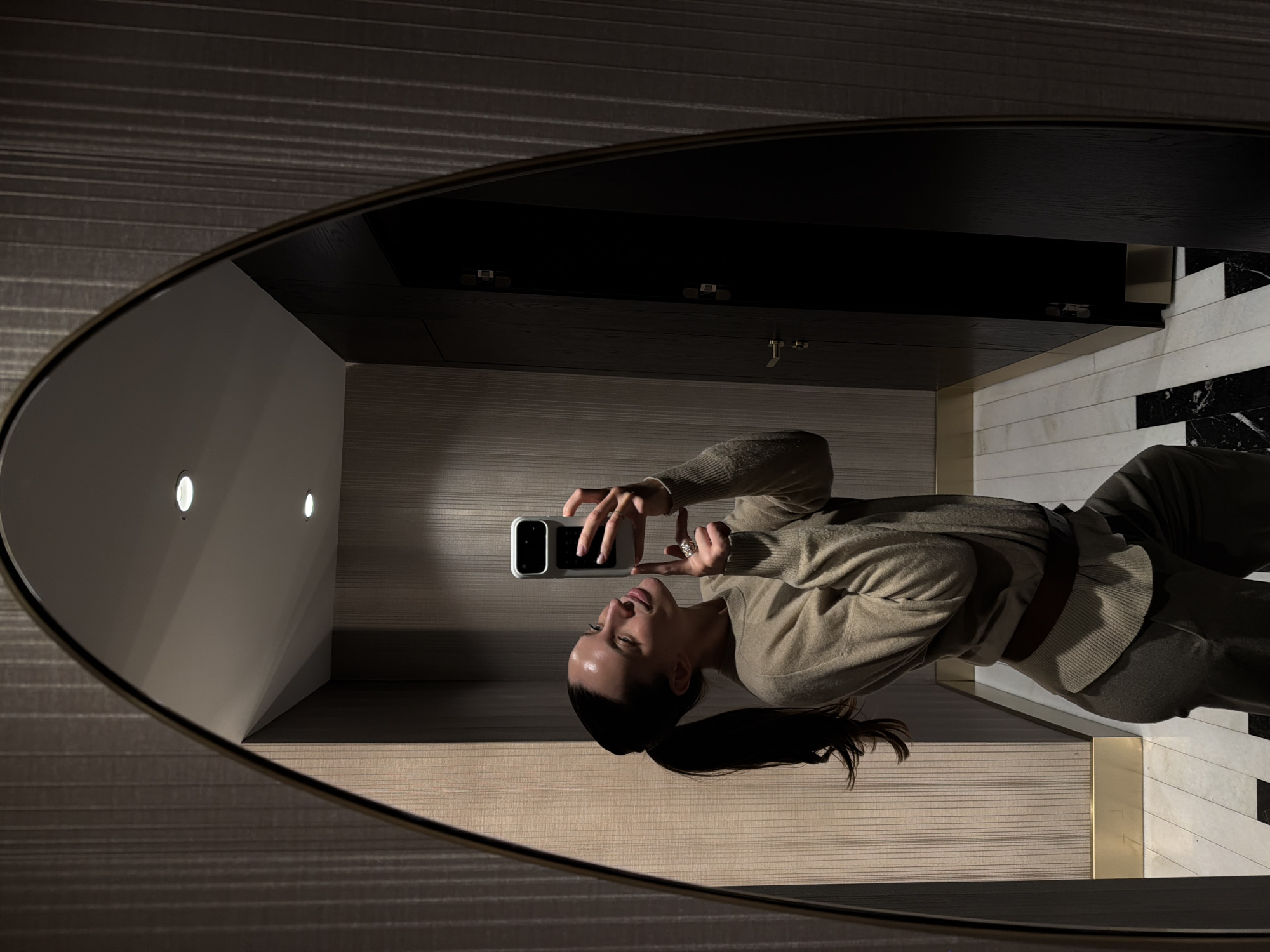 A woman takes a selfie in a mirror. The mirror has an oval shape and a thin dark frame. The woman is wearing a beige sweater and olive-colored pants with a brown belt. Her dark hair is pulled back in a ponytail. She holds a white phone in her left hand and poses with her right hand near her face. The reflection shows part of a room with a light-colored wall, ceiling spotlights, and a dark countertop. The location cannot be accurately determined as Budva, Montenegro, based solely on this image.