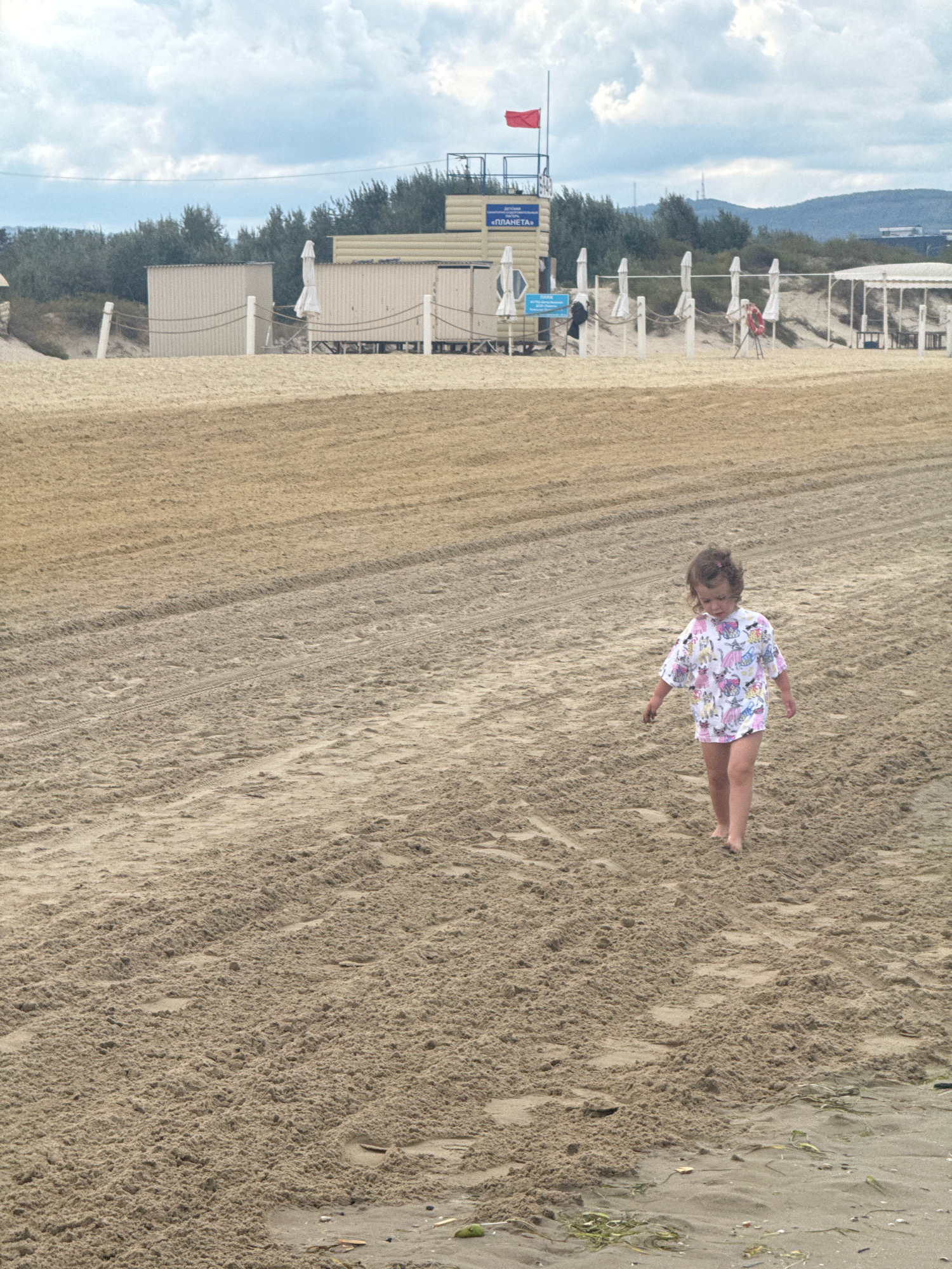 A young child with dark curly hair, wearing a short-sleeved shirt with a colorful pattern, walks barefoot on a sandy beach. The child is facing slightly to the left of the camera. In the background, there are several beach structures, including what appears to be a lifeguard station with a red flag, several covered seating areas with umbrellas, and other buildings. The beach is mostly empty, with tire tracks visible in the sand. The sky is overcast with clouds. The location is the beach in Anapa, Russia.
