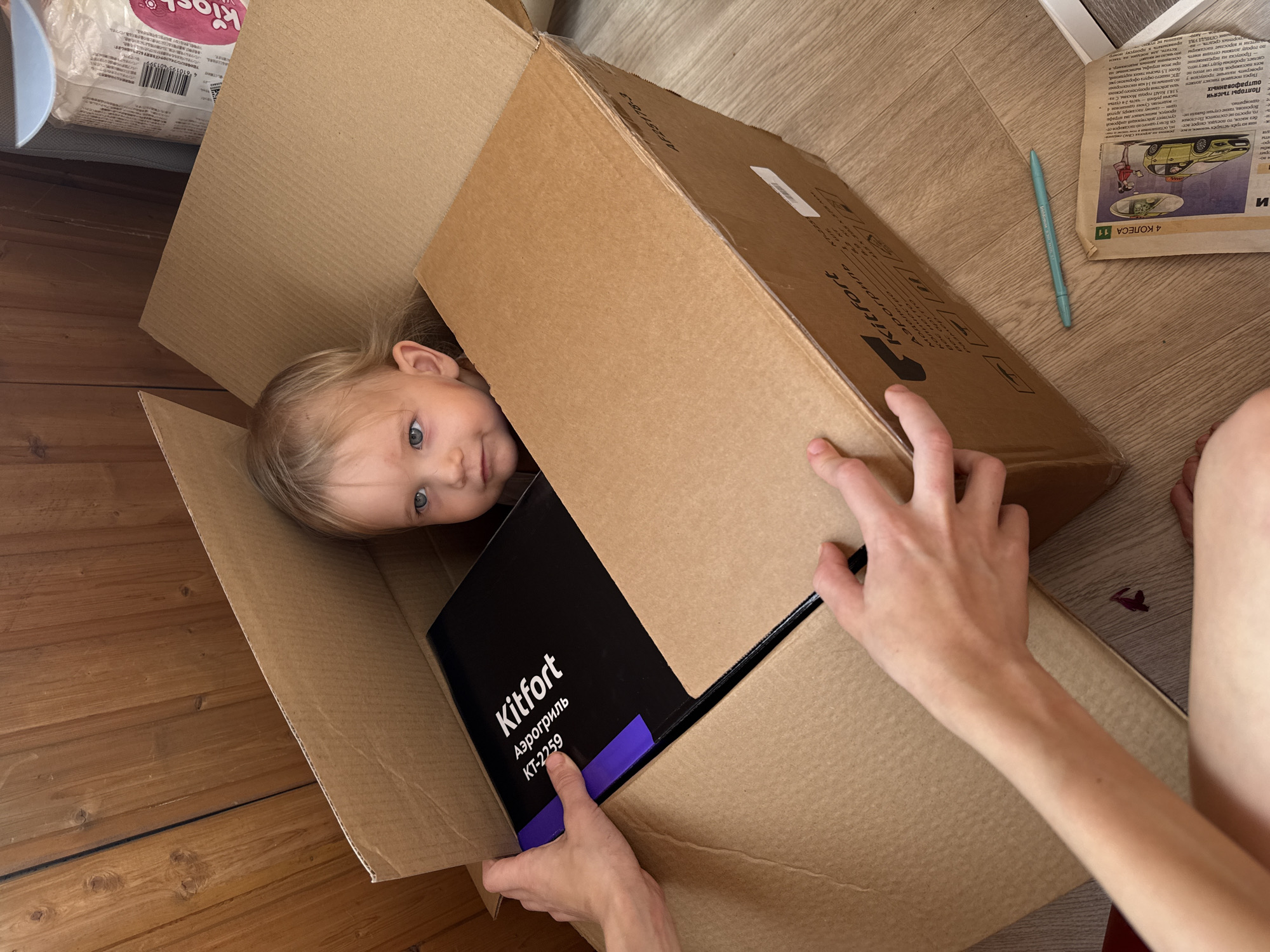 In this overhead shot, a young child with blonde hair and blue eyes peeks out from inside a large cardboard box. The child's head is tilted, and they are looking directly at the camera with a curious expression. The box is open, and a black box with white text is visible inside, along with a purple tab. The cardboard box is placed on a wooden floor, suggesting an indoor setting. A person's hands are visible on the right side of the frame, holding the box or reaching into it.
To the right of the main scene, a portion of a newspaper is visible, with headlines and images, including a cartoon of a car and two figures. Some of the visible text on the newspaper appears to be in Russian, with phrases like "Аэрогриль," "Kitfort," and "4 КОЛЕСА." The newspaper itself seems to be dated around May 14th, with references to traffic regulations and fines in Moscow. A blue pen also lies on the newspaper. The overall scene suggests a casual, perhaps playful, moment as the child explores or plays with the box and its contents. The lighting suggests it is daytime.