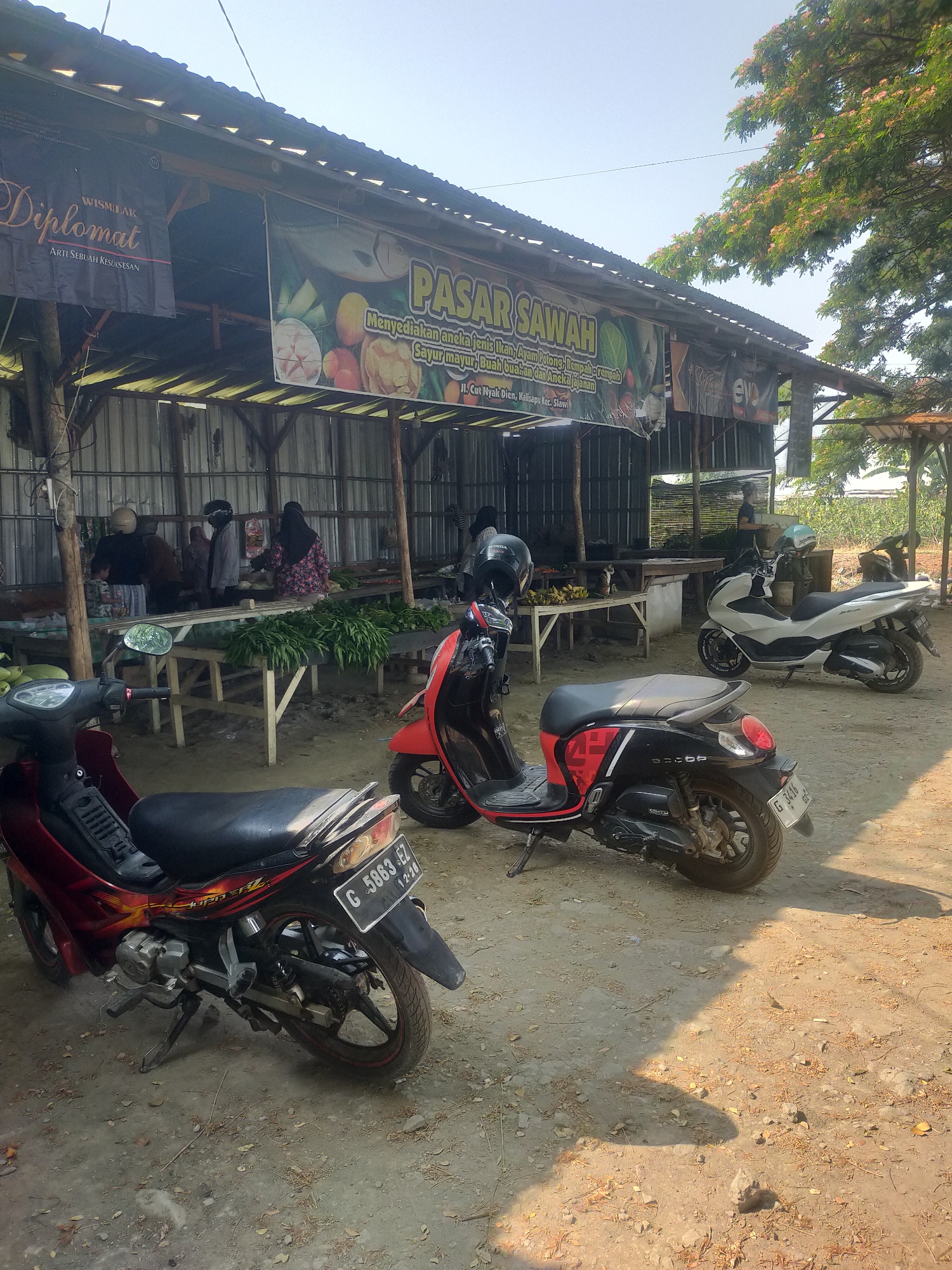 An outdoor market scene is depicted on a sunny day in Kalisapu, Indonesia. The market stalls, constructed with a metal roof and wooden poles, are set up along a dirt ground. A banner hanging from the roof reads "PASAR SAWAH" (Rice Field Market) and advertises "Menyediakan aneka Ikan, Ayam Potong, Rempah-rempah, Sayur mayur, Buah buahan dan Aneka jajanan" (Provides various fish, chicken, spices, vegetables, fruits, and snacks). Another banner to the left says "WISMA LAK Diplomat ARTI SEBUAH KESUKSESAN" (Wisma Lak Diplomat, The Meaning of Success).
Several motorcycles are parked in front of the stalls. In the foreground, a red and black scooter is parked facing right, with its license plate partially visible. To its left, a red motorcycle with a black seat is parked facing left. Further back, a white scooter is parked facing right, and another motorcycle is visible in the far background.
Inside the market, several people are seen behind the stalls, interacting with each other or tending to the produce. Tables are laden with fresh vegetables, including large bunches of green leafy vegetables, and some fruits like bananas are also visible. The overall impression is a local market providing fresh produce and goods to the community.