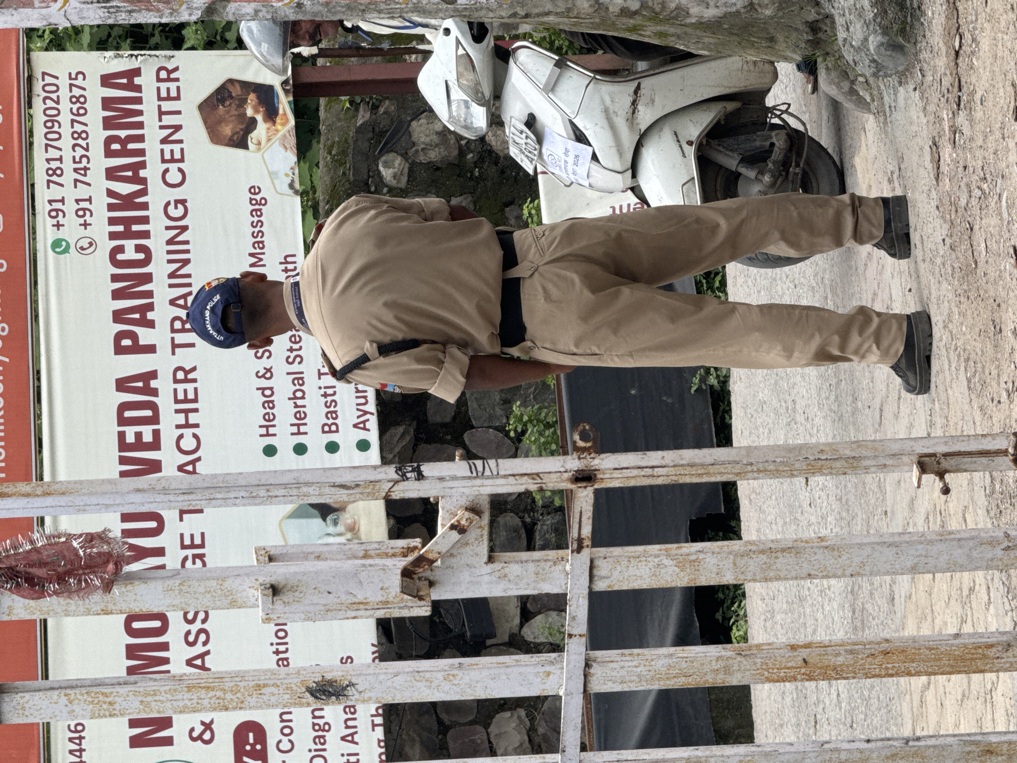 The image depicts an outdoor scene in Rishikesh, India, likely near a training center. In the foreground, a white metal gate with visible rust and peeling paint stands partially open. Behind the gate, a uniformed police officer from Uttarakhand is facing away from the camera, wearing a tan uniform and a cap with "UTTARAKHAND POLICE" visible. The officer appears to be standing guard or observing something.
To the right of the officer, a white scooter is parked. A sign behind the officer advertises "N MO YU VEDA PANCHKARMA TEACHER TRAINING CENTER," listing services such as "Head & S Massage," "Herbal Ste ath," "Basti T Ayur." Phone numbers and a logo are also visible on the sign. The environment suggests a somewhat rural or roadside setting, with stone walls and greenery in the background. The lighting indicates it is daytime.
