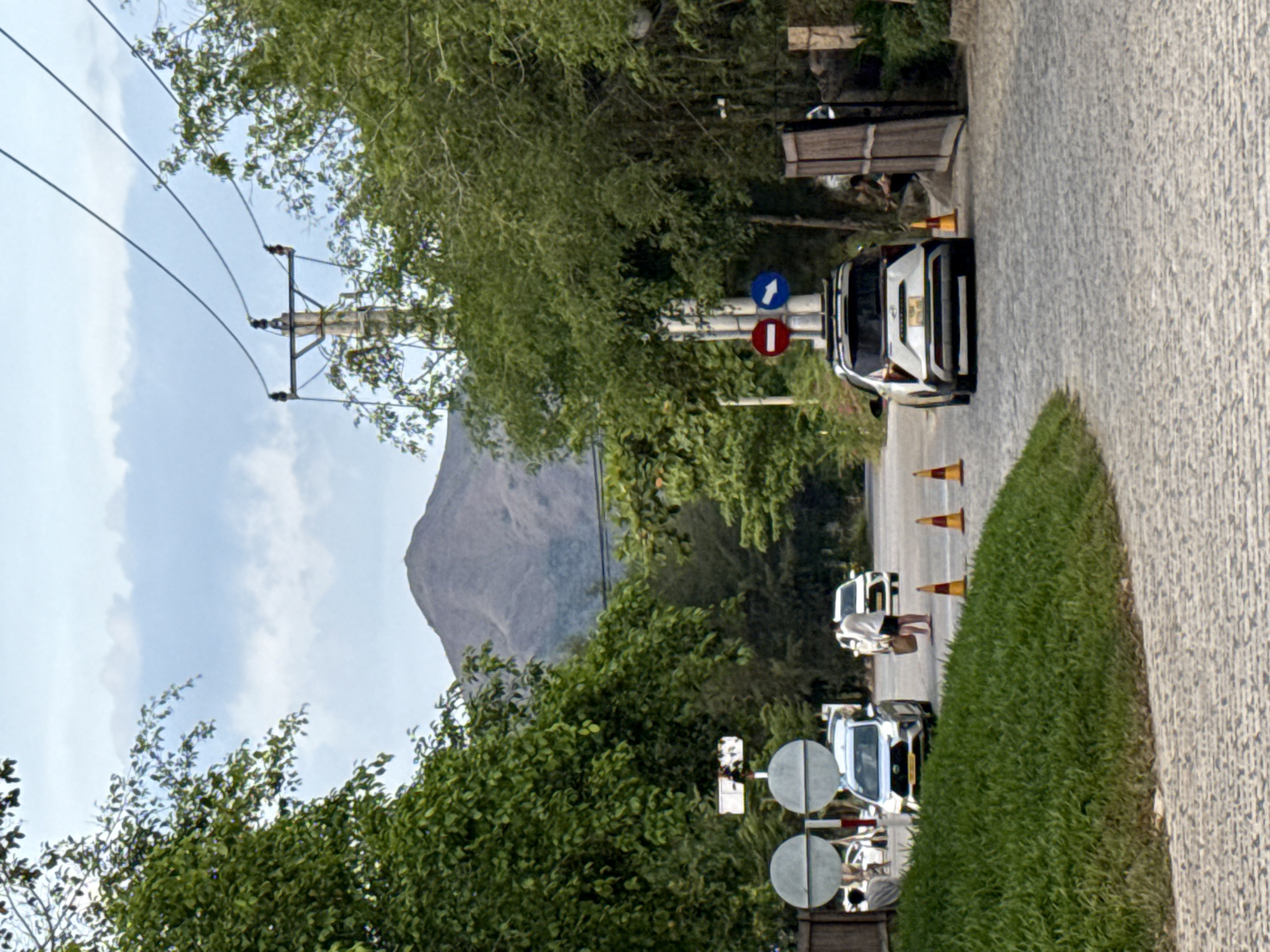 The image depicts a street scene in Tay Nha Trang Ward, Vietnam. A mountain rises in the background. The street is paved with light-colored stones and is bordered by a green grassy area on one side. Several white vehicles are parked along the street, accompanied by orange traffic cones. One of the vehicles has a sign with traffic directions. A power line pole is visible on the left side of the frame. Trees and shrubbery are interspersed along the roadside. People are present near the parked vehicles, although their actions are not readily discernible.