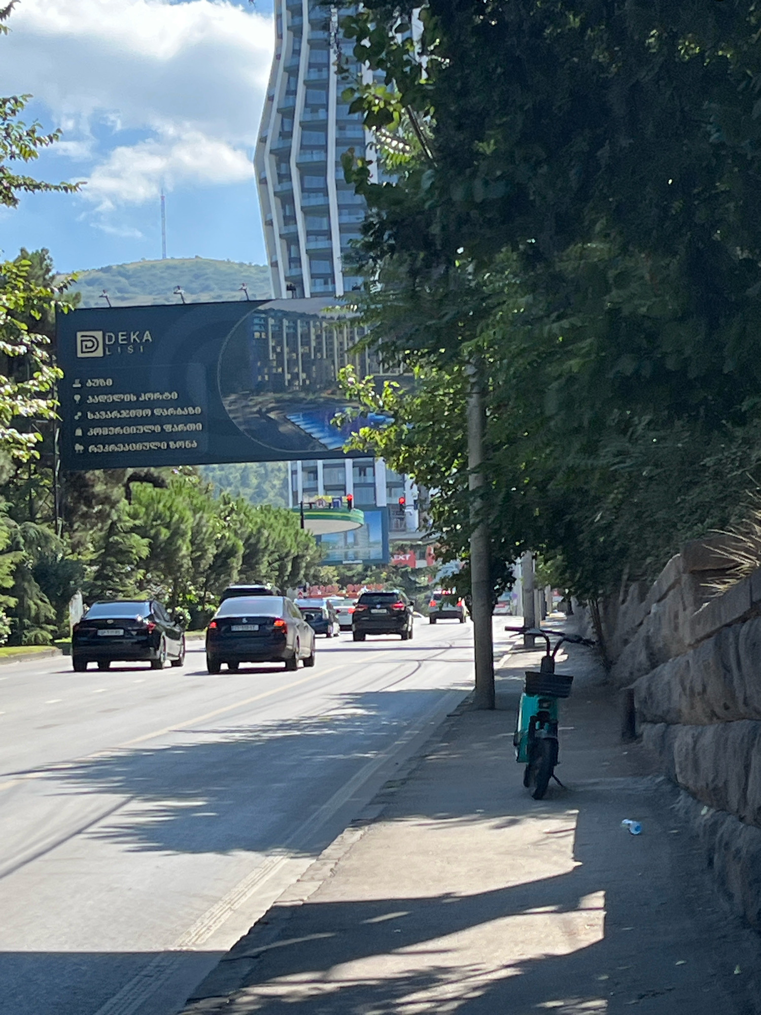 The image captures a street scene in Tbilisi, Georgia. A modern, tall building with a distinctive architectural design stands in the background, partially obscured by lush green trees. A large billboard advertising "DEKA LIST" is prominently displayed, featuring images of apartments and amenities. A multi-lane road runs through the foreground, with several dark-colored cars driving on it. A turquoise electric scooter is parked on the sidewalk to the right, next to a stone wall. The sunlight casts long shadows across the road and sidewalk, indicating it is likely late afternoon. The sky is partly cloudy with a visible cell tower on a distant hill.