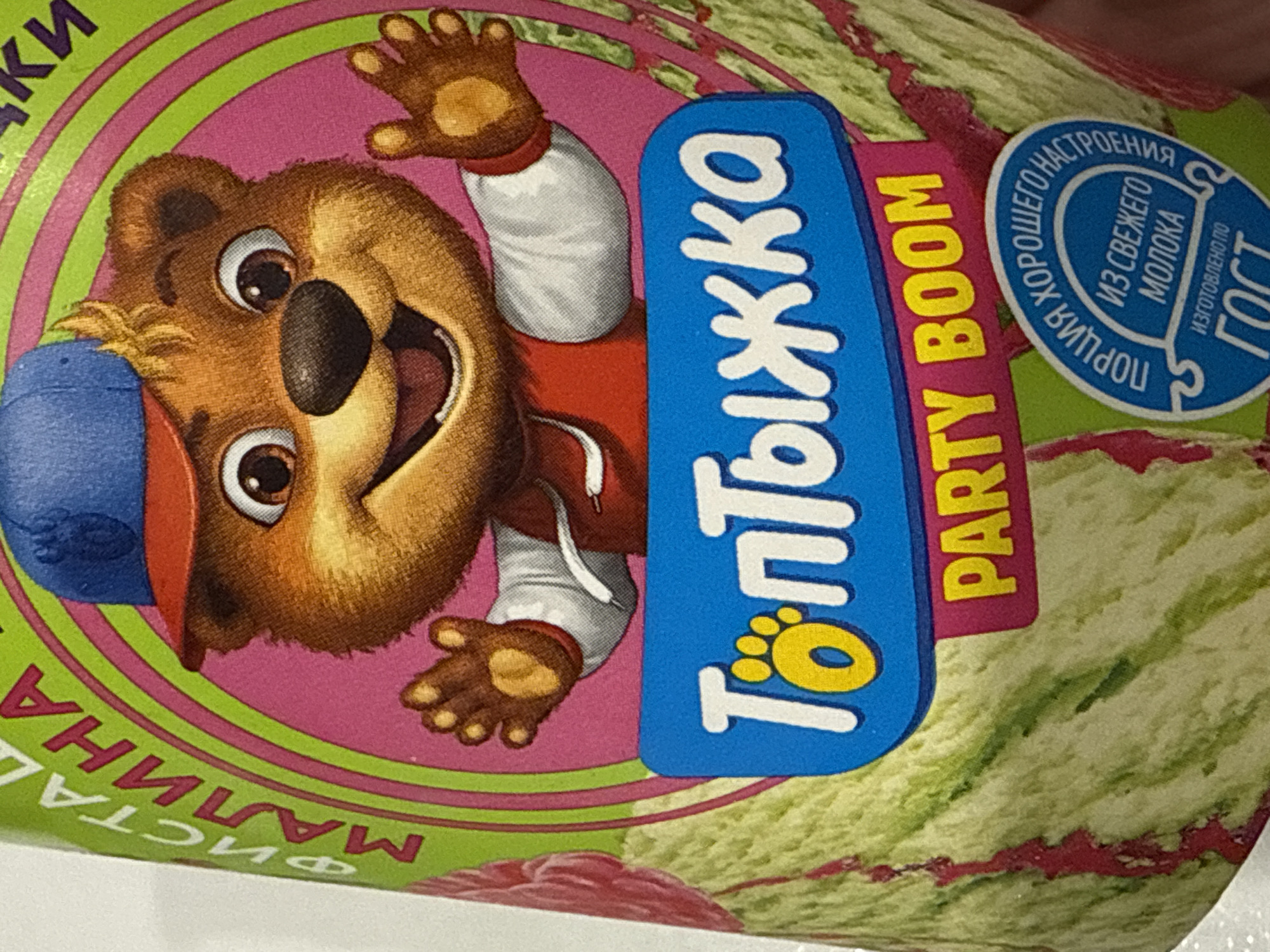 This is a close-up shot of a container featuring a cartoon bear character wearing a red hooded sweatshirt and a blue baseball cap. The bear is depicted with outstretched arms against a pink background with green concentric circles. A blue emblem with white Cyrillic text reads "Топтыжка" (Toptyzhka), with a yellow paw print within one of the letters. Adjacent to the emblem is a pink banner displaying the words "PARTY BOOM" in yellow. In the upper right corner, there's a blue circular emblem containing Cyrillic text that includes phrases indicating "from fresh milk" and a quality standard or regulation "ГОСТ" (GOST). The container itself has a mottled green and red pattern, reminiscent of a watermelon rind. Cyrillic text that reads "ФИСТА МАЛИНА" is visible on the left side of the image. This is likely the labeling of a food product, possibly ice cream, by the company Toptyzhka. There is no reference to the city of Votkinsk, Russia, in the image.