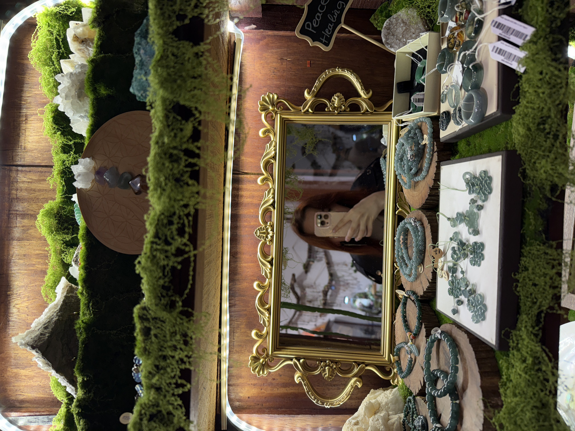 This media file captures an intricately arranged indoor display, likely from a shop or market stall, showcasing a variety of crystals, natural stones, and jewelry. The image is oriented vertically but depicts a horizontal display, with warm wooden shelving forming the background, extensively adorned with vibrant green moss, giving the scene a natural and earthy aesthetic.
On the left side of the display, large, clear and white crystal clusters are prominent, nestled within the moss. Below them, a circular wooden disc engraved with a "Flower of Life" pattern holds smaller, polished stones including amethyst and other colorful tumble stones. A strip of white LED lighting runs along the top-left edge, illuminating this section.
Centrally positioned is a small, ornate mirror with a golden, rococo-style frame. Reflected in the mirror is the person taking the photograph, showing their reddish-brown hair and a light-colored smartphone with multiple cameras. Above the mirror, a small chalkboard-style sign on a stick reads "Peace" and "Healing" in white script.
The right side of the display features numerous pieces of jewelry, predominantly made from greyish-green stones, possibly jade or a similar material. These include beaded bracelets displayed on small wooden slices, and rings and carved pendants presented on white display boards. Some items have small price tags attached, though the text is illegible. A large, rough crystal or geode is also visible among the green jewelry.
The overall scene suggests a serene and mindful setting, focusing on natural beauty and possibly spiritual or wellness themes. The lighting seems to be a mix of warm artificial light from the LEDs and possibly ambient indoor light, creating a cozy atmosphere. No specific time of day or outdoor weather is discernible. The image lacks direct cues for Da Lat, Vietnam, beyond the user-provided context.