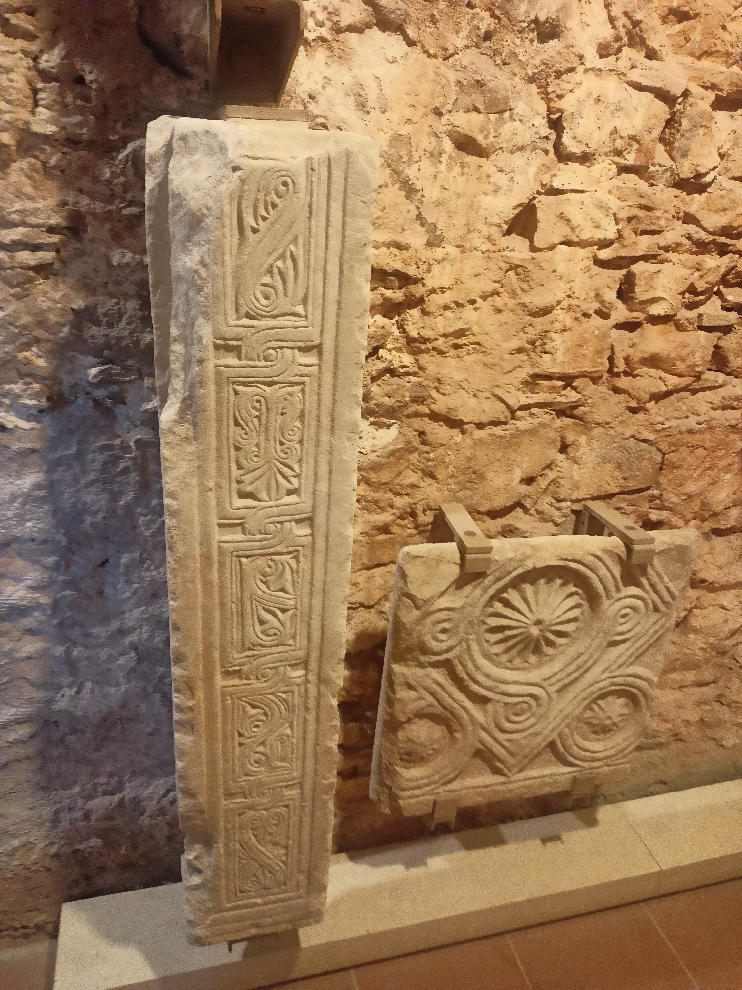 In the Municipal Unit of Anthidon, Greece, an archeological display features two stone artifacts against a rough, tan stone wall. The artifact on the left is a tall, rectangular stone pillar, standing upright. Its surface is divided into rectangular panels, each containing intricate carvings of scrolling patterns and stylized plant-like motifs. The artifact on the right is a smaller, squarish stone block, positioned horizontally. It is adorned with a central circular design resembling a flower or starburst, surrounded by swirling, curvilinear patterns. This artifact is held in place by two light-colored, angled brackets. The floor in front of the artifacts appears to be tiled in a reddish-brown color. The lighting suggests an indoor environment, possibly a museum or exhibition space.