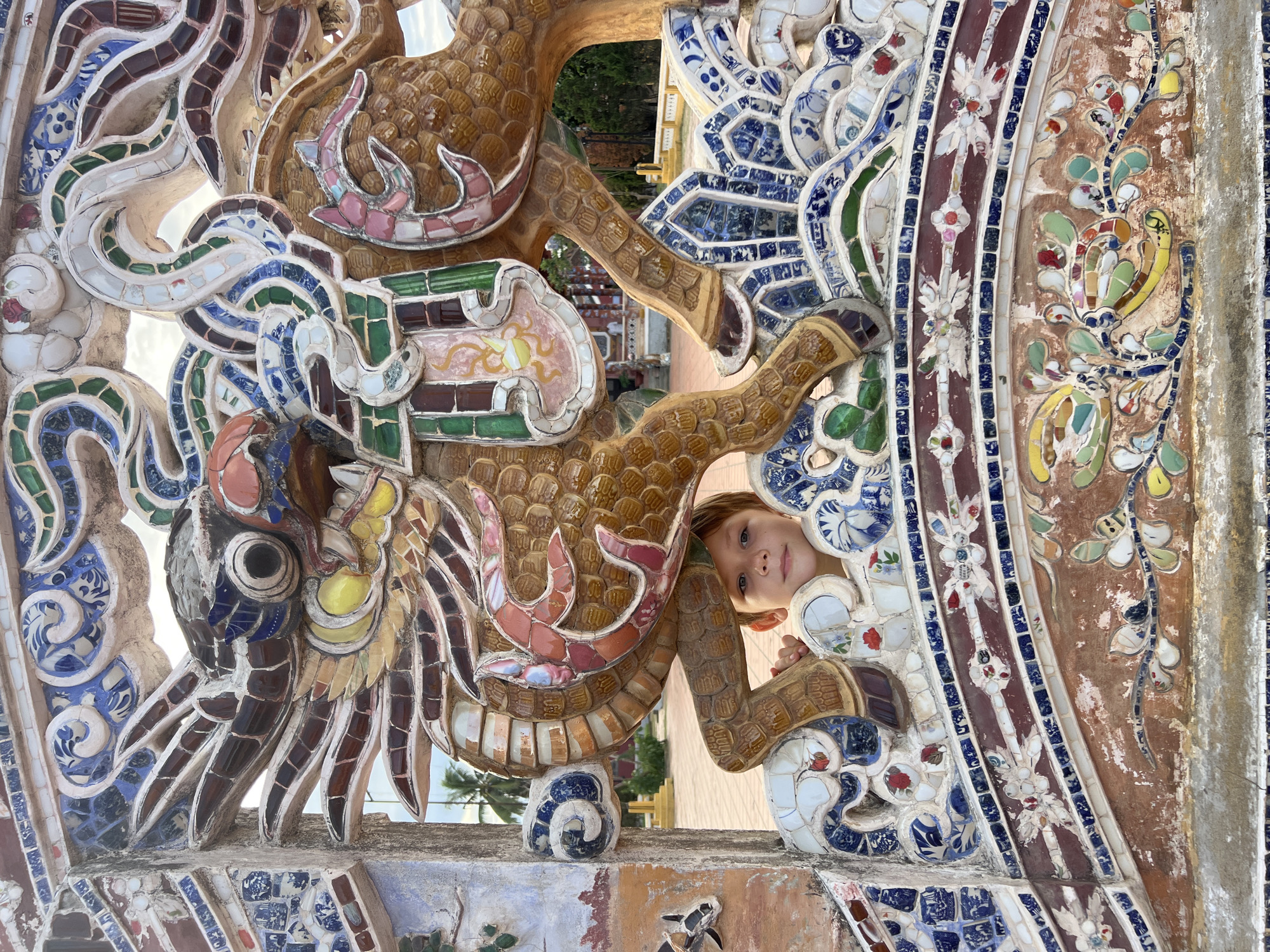 This is a close-up, eye-level, full shot of a young boy peeking through an elaborately decorated mosaic structure, likely at the Tomb of Khai Dinh in Hue, Vietnam, not Hoi An. The structure is a detailed mosaic sculpture of a dragon, with a brown body and intricate blue, white, green, and red tile work. The boy, with light skin and light brown hair, is smiling and looking directly at the camera. His head and shoulders are visible between the dragon's legs and body. The background shows a slightly blurred view of a courtyard with palm trees and traditional architecture, under a bright sky. The mosaic is made of many small, colorful ceramic pieces, forming patterns of clouds, waves, and floral designs. The boy's left hand is visible, touching the mosaic.