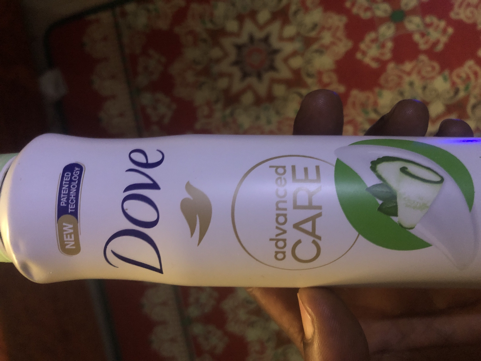 A person's hand holds a white Dove Advanced Care deodorant stick. The deodorant has a green band around the bottom with images of cucumber slices and leaves. The background is a red and yellow patterned cloth. The image was taken in Kabong, Nigeria.