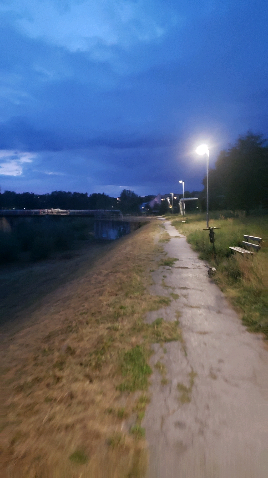 The image captures a twilight scene along a paved pathway next to a body of water, likely a dam or reservoir, in Prešov, Slovakia. The sky is a deep, moody blue with dramatic clouds, suggesting an approaching storm or the aftermath of one. A single lamppost illuminates the pathway, casting a bright glow. To the right of the path, a wooden bench and a parked scooter are visible amidst overgrown grass. In the distance, trees and some buildings can be seen on the horizon. The water on the left reflects the dusky sky. The overall impression is one of a tranquil, yet slightly ominous, natural setting within an urban or suburban environment.