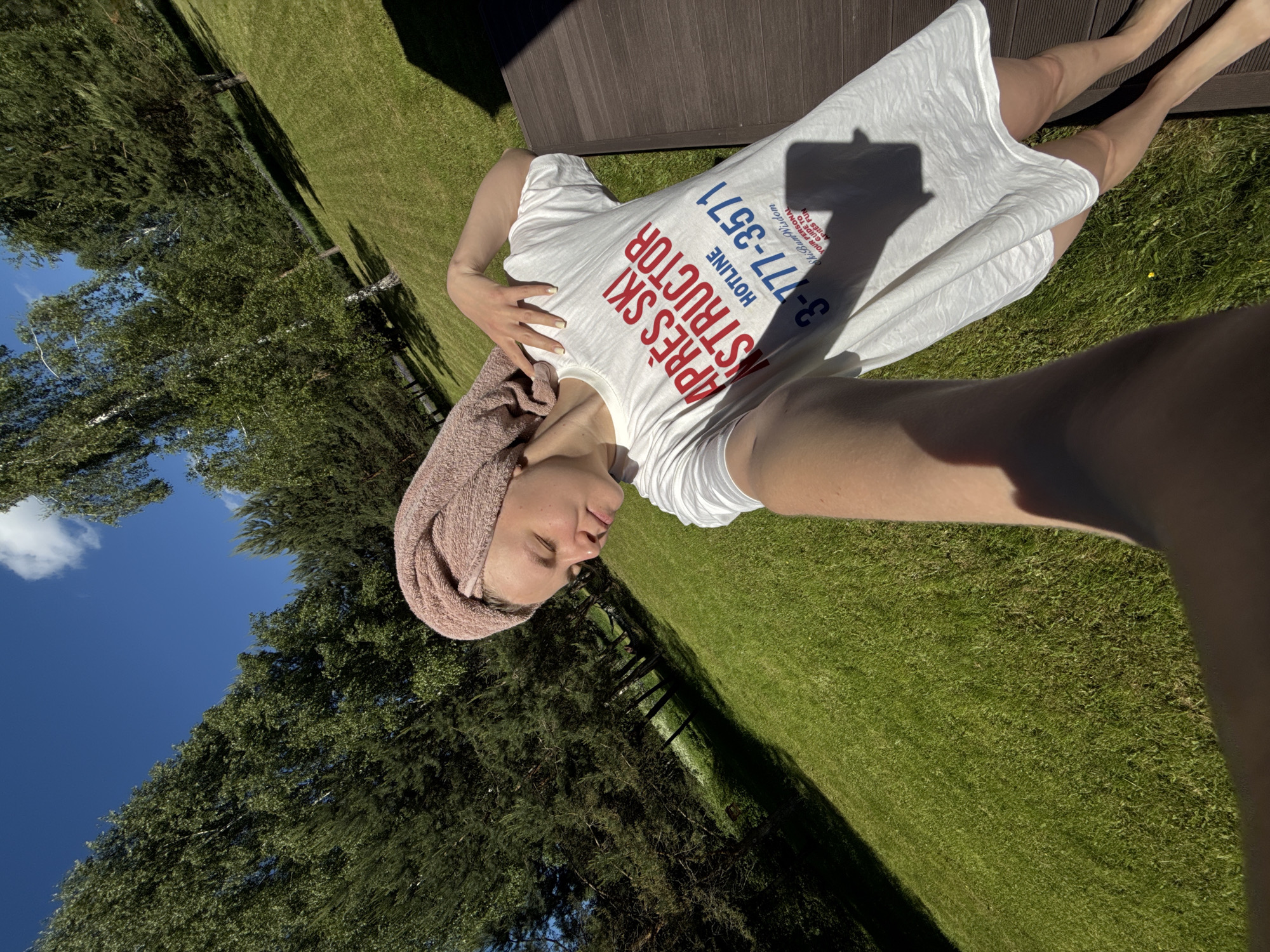 An outdoor selfie depicts a person, appearing female, situated on a dark brown slatted wooden surface in Копнино, Russia. The individual has a light pink towel wrapped around their head and wears an oversized white t-shirt. The t-shirt prominently displays "APRÈS SKI INSTRUCTOR" in red text, "HOTLINE 3-777-3571" in blue, and smaller text reading "SkiBum Wisdom YOUR PERSONAL GUIDE TO APRÈS FUN". The person's left hand, adorned with light-colored nail polish, rests on their neck. Their head is tilted, and eyes are partially closed. Bare legs and feet are visible on the right. The background features an expansive, brightly sunlit green lawn, bordered by a dense line of deciduous and coniferous trees casting long shadows. Above the tree line, a clear blue sky with sparse white clouds is visible.