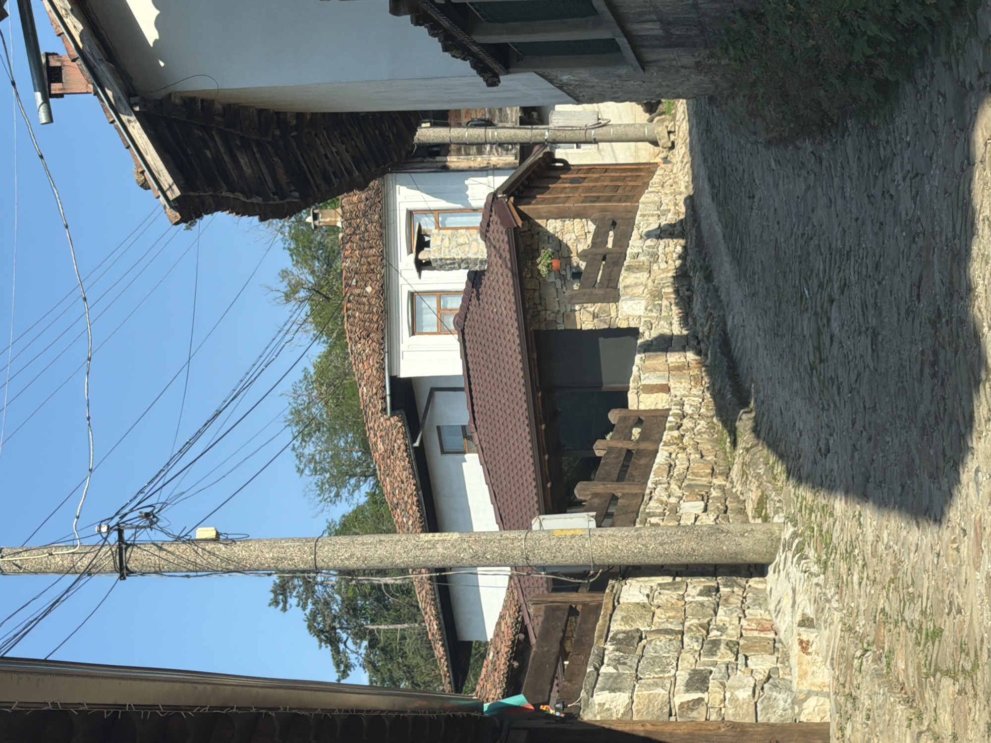 The image depicts a static, outdoor scene of a traditional cobblestone street winding uphill in Elena, Bulgaria, under a clear, bright blue sky. No individuals or animals are present.
In the foreground, an uneven cobblestone street, predominantly grey and tan with sparse green vegetation growing between stones, curves gently to the right and ascends. A distinct, elongated shadow from an unseen object extends diagonally from the lower left to the mid-right of the street. On the left side of the street, a rustic stone wall, constructed from varied light-colored rocks, supports a wooden fence or railing. A dark wooden post is partially visible on the far left edge. Centrally positioned, a tall, round, grey utility pole, likely concrete, rises, heavily laden with multiple black and a few white electrical wires that stretch across the blue sky. Several small, rectangular electrical boxes are affixed to the pole. On the right, the lower portion of another stone wall, partially covered with small green plants, frames the right edge of the street.
In the midground, several traditional Bulgarian houses are visible. The closest structure on the left features a brown tiled roof and a stone-clad lower facade with a small wooden railing or balcony. A single terracotta pot with a green plant is affixed to the stone wall. Further back and to the right, a prominent white-walled house with a red-brown tiled roof and dark wooden eaves has multiple windows with wooden frames and a distinct stone chimney extending above the roofline. Another house with similar architectural features is partially visible behind it. Exposed wooden beams are noticeable under the eaves of some roofs.
The background consists of a vibrant, cloudless blue sky, indicating fair weather, and a dense cluster of green trees, including tall conifers and deciduous trees, suggesting the proximity of forested hillsides. The overall lighting is bright, casting sharp shadows, consistent with direct sunlight.