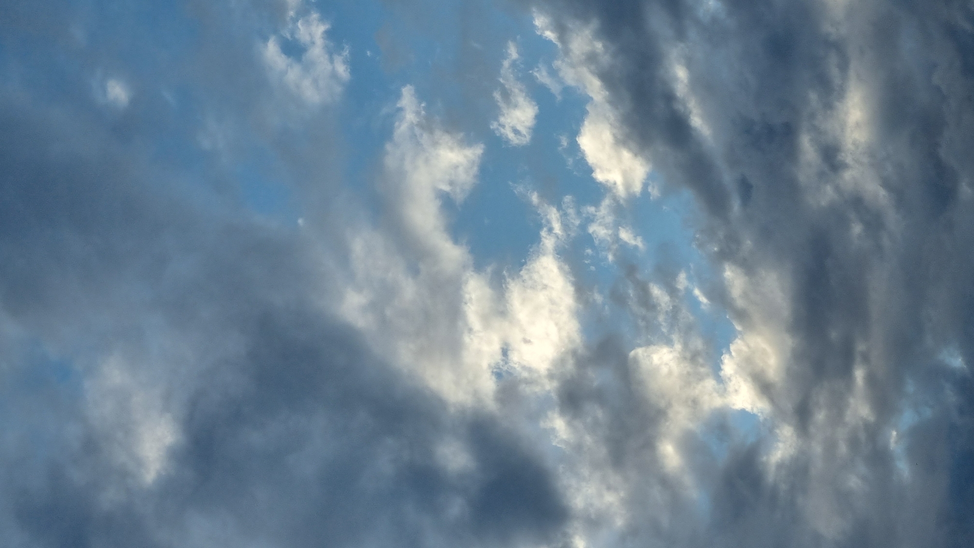 The image shows a sky filled with a mix of dark and light gray clouds, with patches of blue sky visible between them. The lighting suggests it is either early morning or late afternoon. There are no people, subjects, or objects visible in the frame, only the natural sky. The location in Vysokoe, Russia, is not discernible from the image itself.