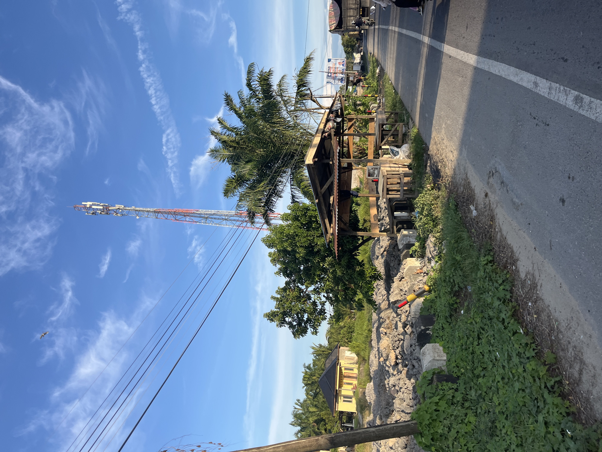 In Seuneubokpunti, Indonesia, a rural roadside scene unfolds under a bright blue sky with scattered white clouds. A tall, red and white cellular tower dominates the upper center of the frame, with multiple antennas visible. Several power lines stretch across the sky from left to right. To the left of the tower, a small yellow house with a dark roof is nestled among lush green trees and palm trees. In the foreground, a weathered wooden stall with a corrugated metal roof sits beside the asphalt road, its shelves sparsely stocked. Piles of rocks and concrete blocks are scattered to the left of the stall, with green vegetation growing over some of them. The road features a white dividing line and shows signs of wear. A motorcycle is seen riding on the right side of the road, with a truck in the distance. A single bird is visible soaring high in the sky.